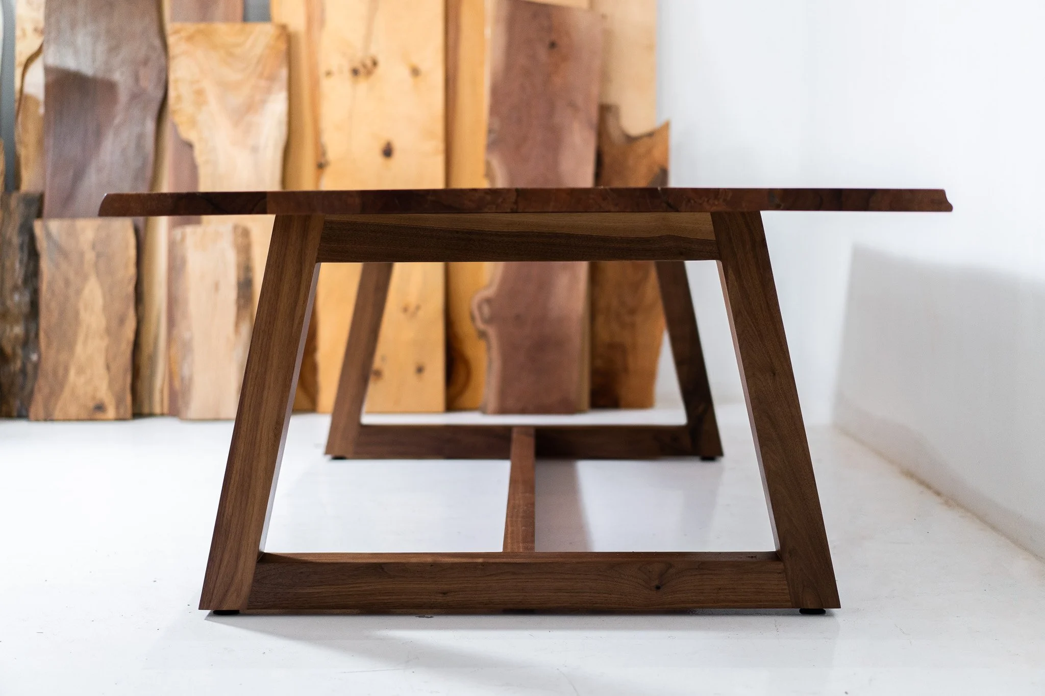 Two wooden tables with angled legs, one in the foreground and one in the background, placed on a white floor with wood panels leaning against the wall behind them.