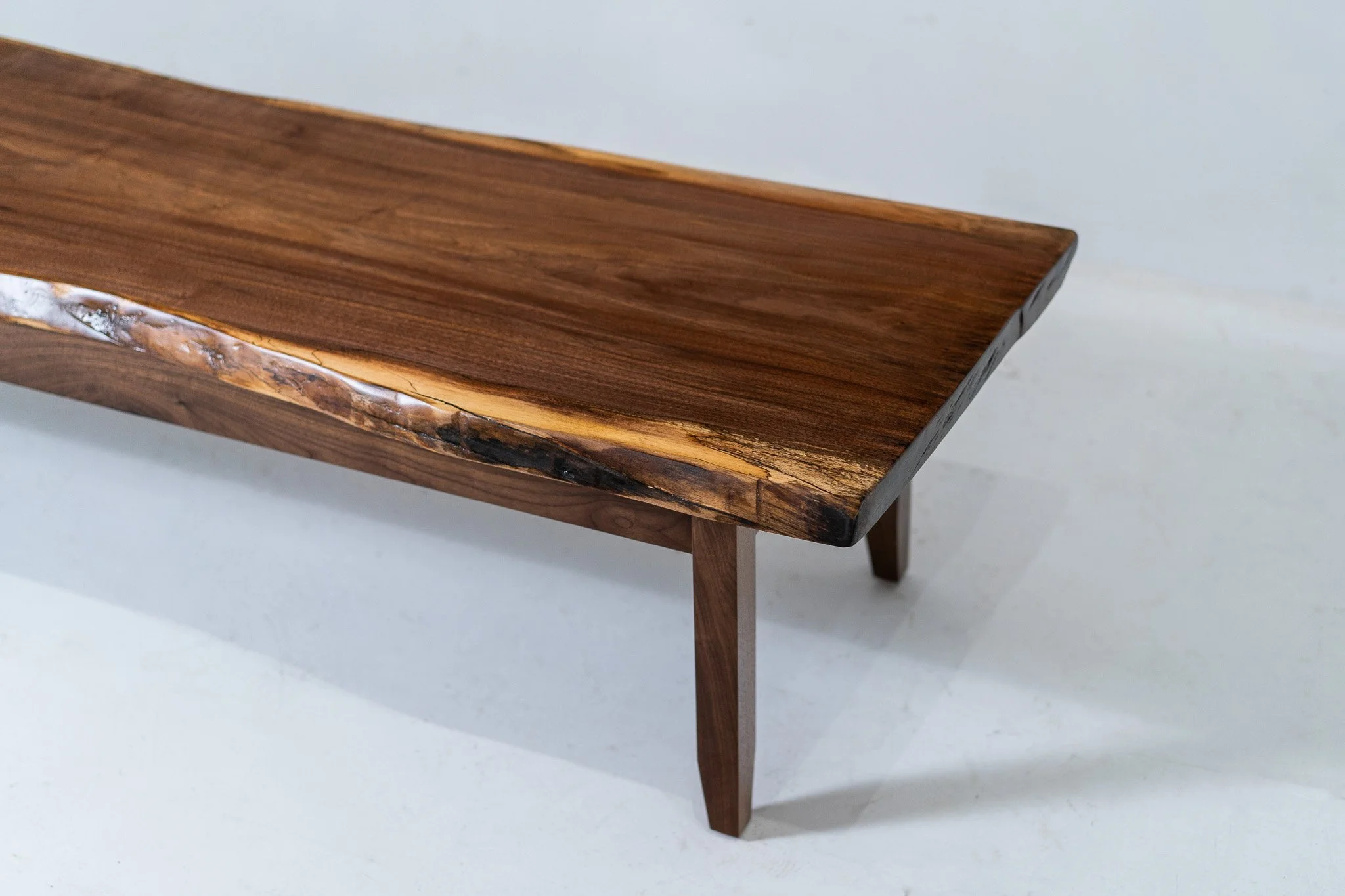 Close-up of a wooden coffee table with a top made of smooth, polished dark wood and a natural edge detail, supported by four slim matching wooden legs, set against a light background.