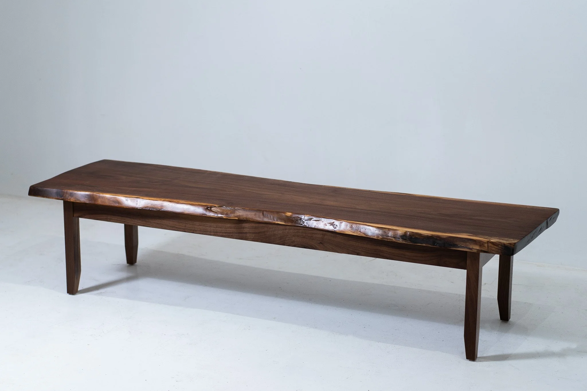 A wooden bench with a natural finish, featuring a live edge on the seat, set against a plain light-colored wall.