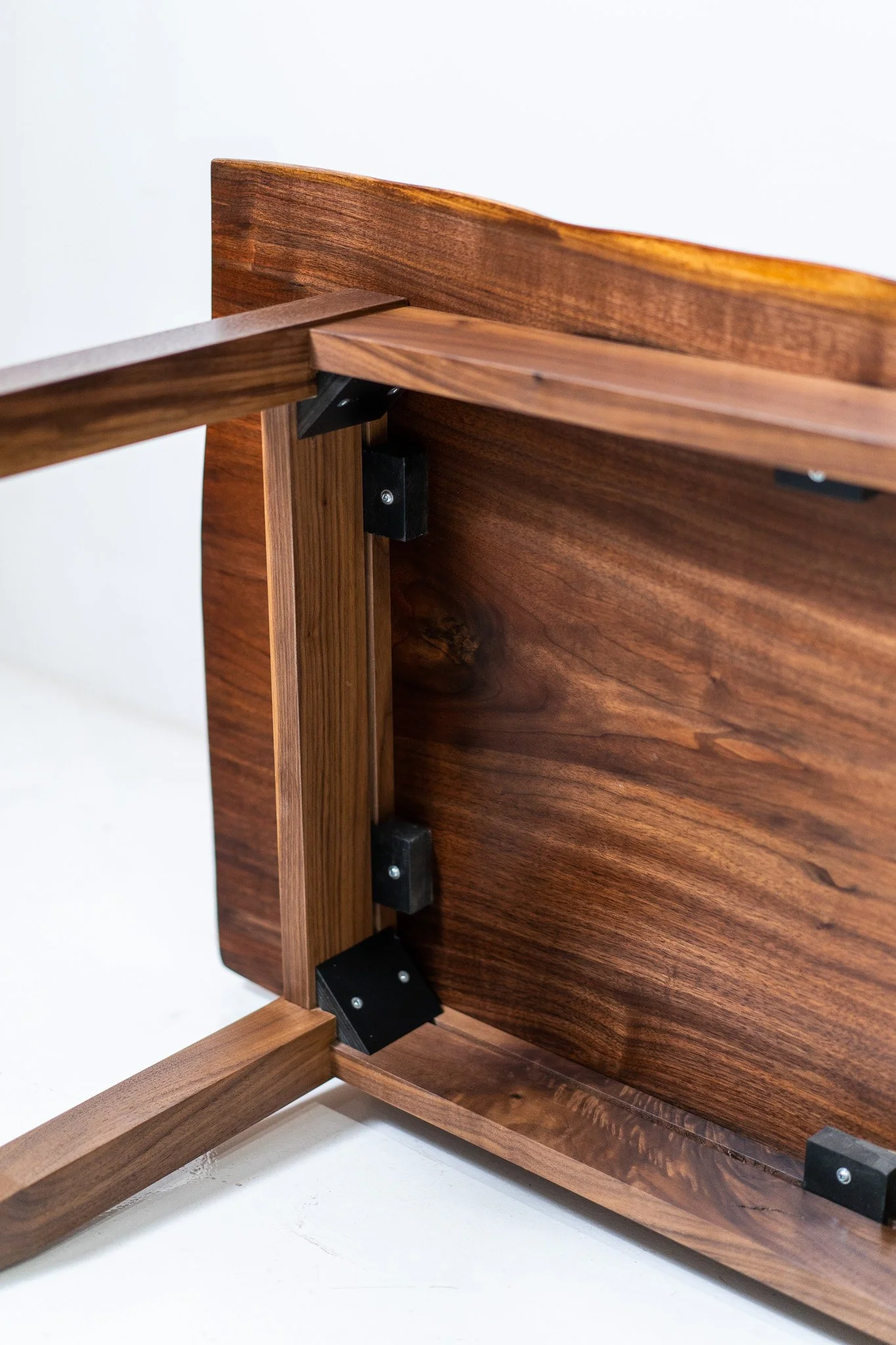 Close-up of the underside of a wooden table with wooden and metal brackets connecting the table leg to the tabletop, showing the wood grain and screws.