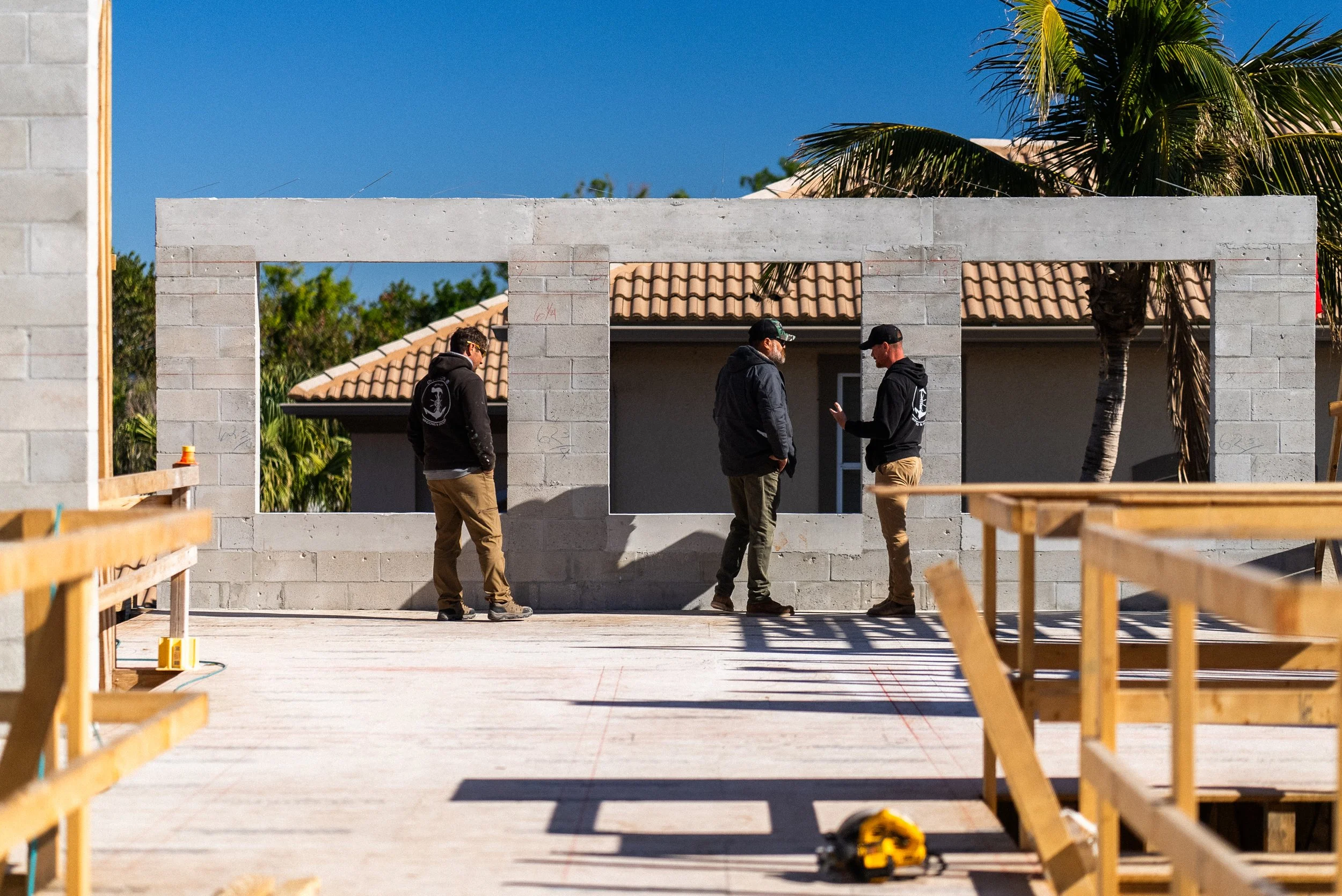 Three construction workers standing and talking on a building site, with a partly built concrete wall and orange tiled roof house in the background, under a clear blue sky.