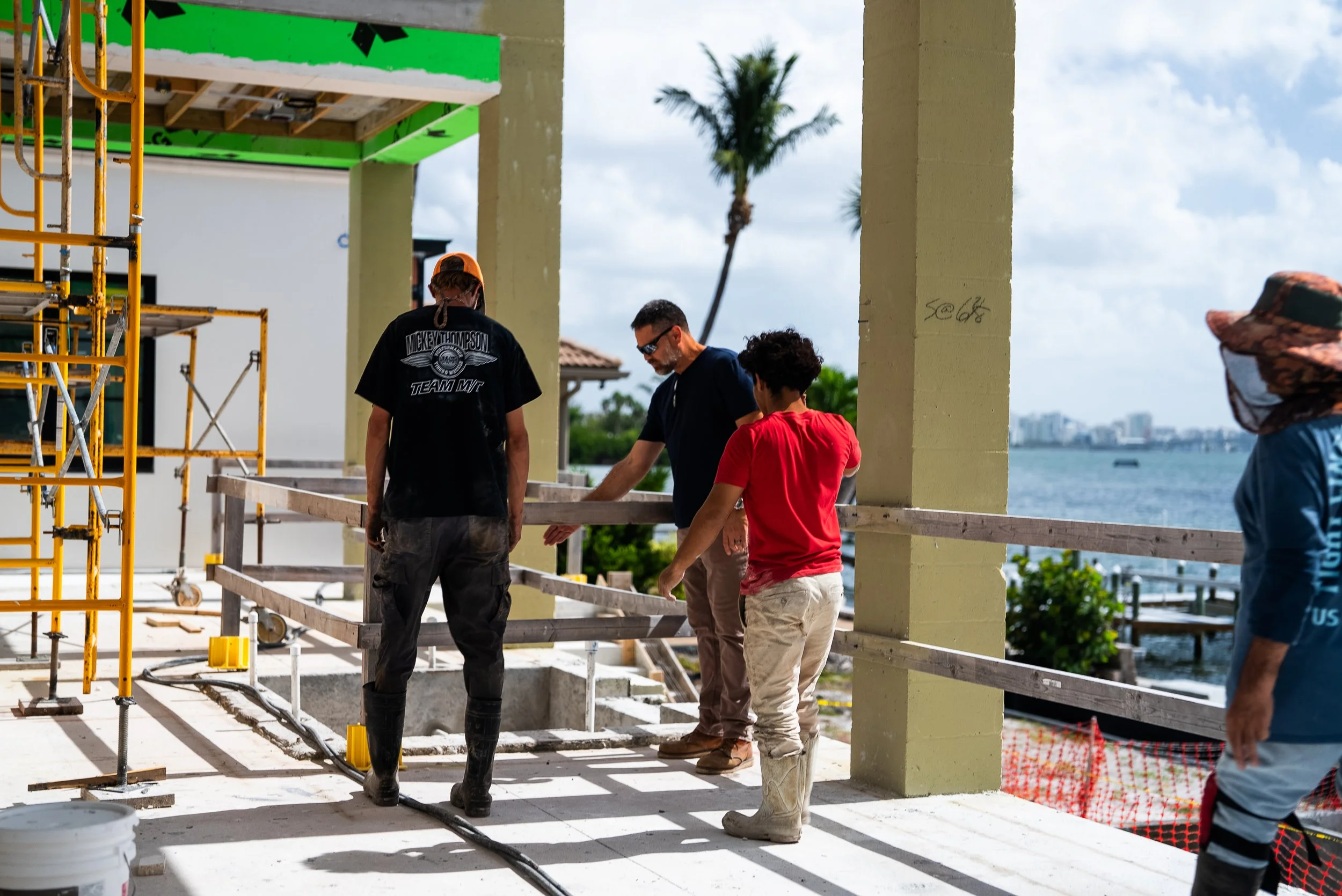 Four construction workers are on a construction site near the waterfront. They are inspecting or discussing a wooden beam. One worker is wearing a Harley-Davidson t-shirt and rain boots, another is in black shirt and sunglasses, the third is in a red