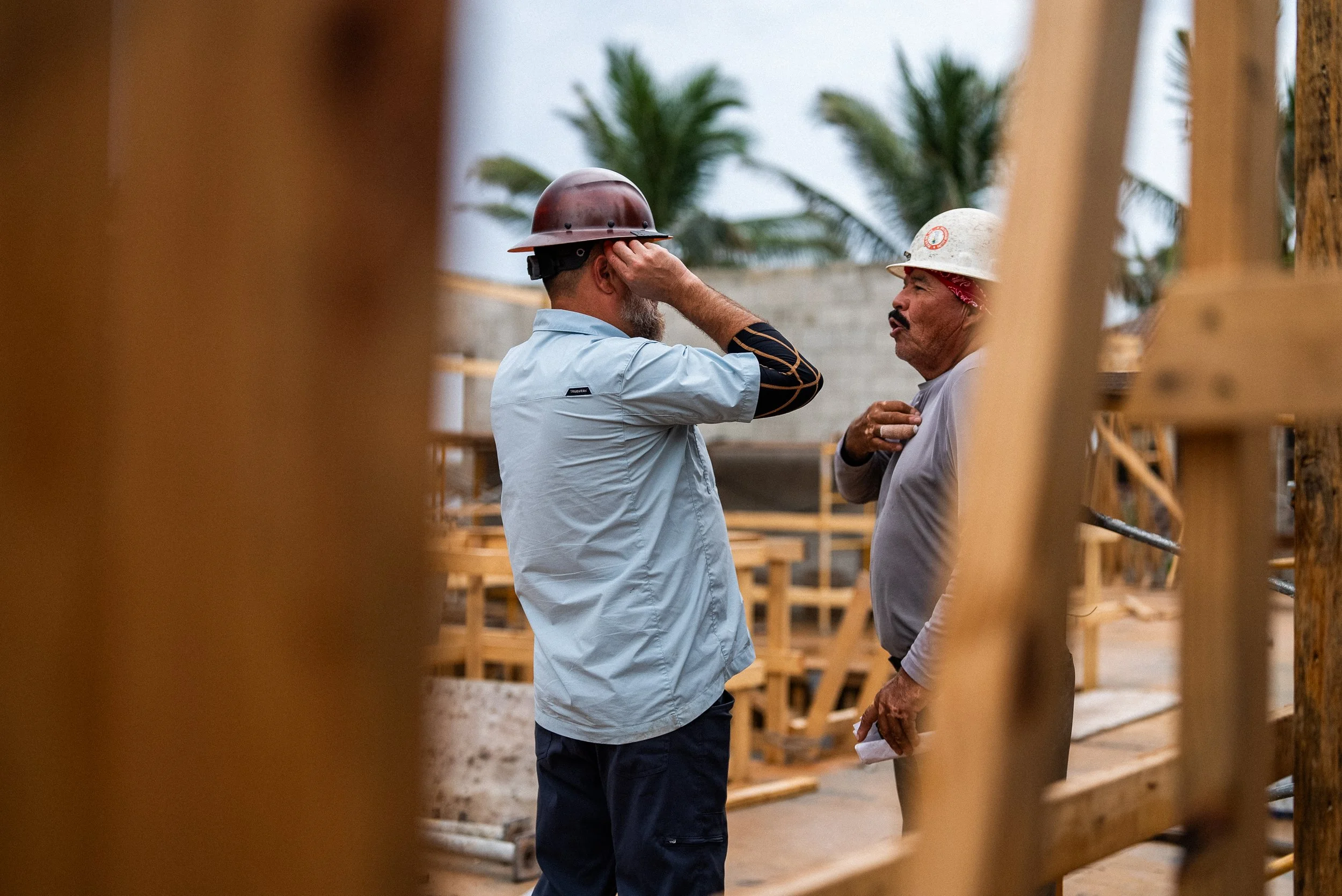 Two construction workers wearing safety helmets and casual work clothes are having a conversation on a construction site. One man is touching his helmet, and the other has his hand on his chest, holding a rolled-up paper. The site has wooden scaffold