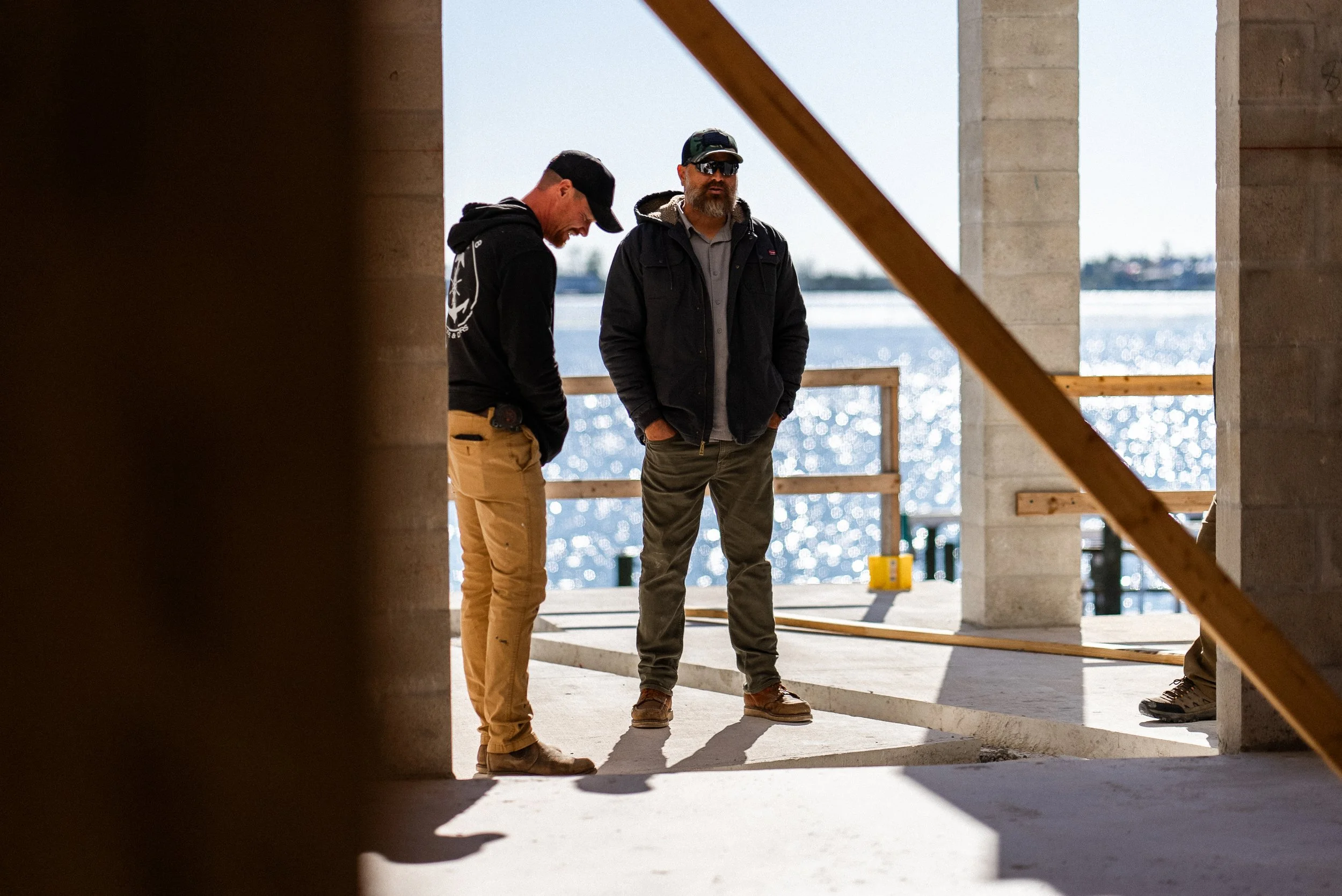 Two construction workers stand on a partially completed outdoor structure near a body of water with sunlight reflecting off it.