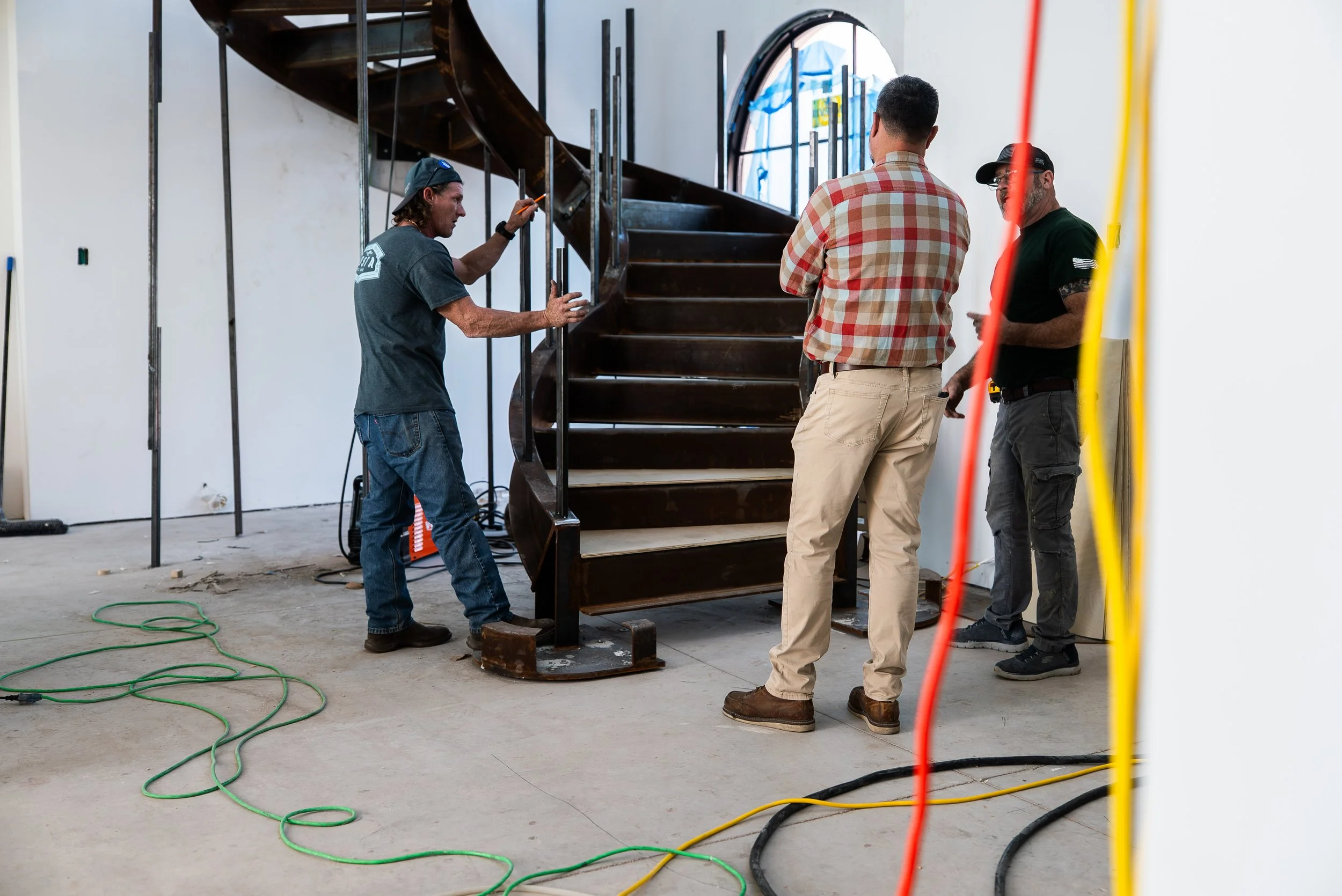 Three men working on a wooden spiral staircase inside a building under construction or renovation, with tools and electrical cords on the floor.