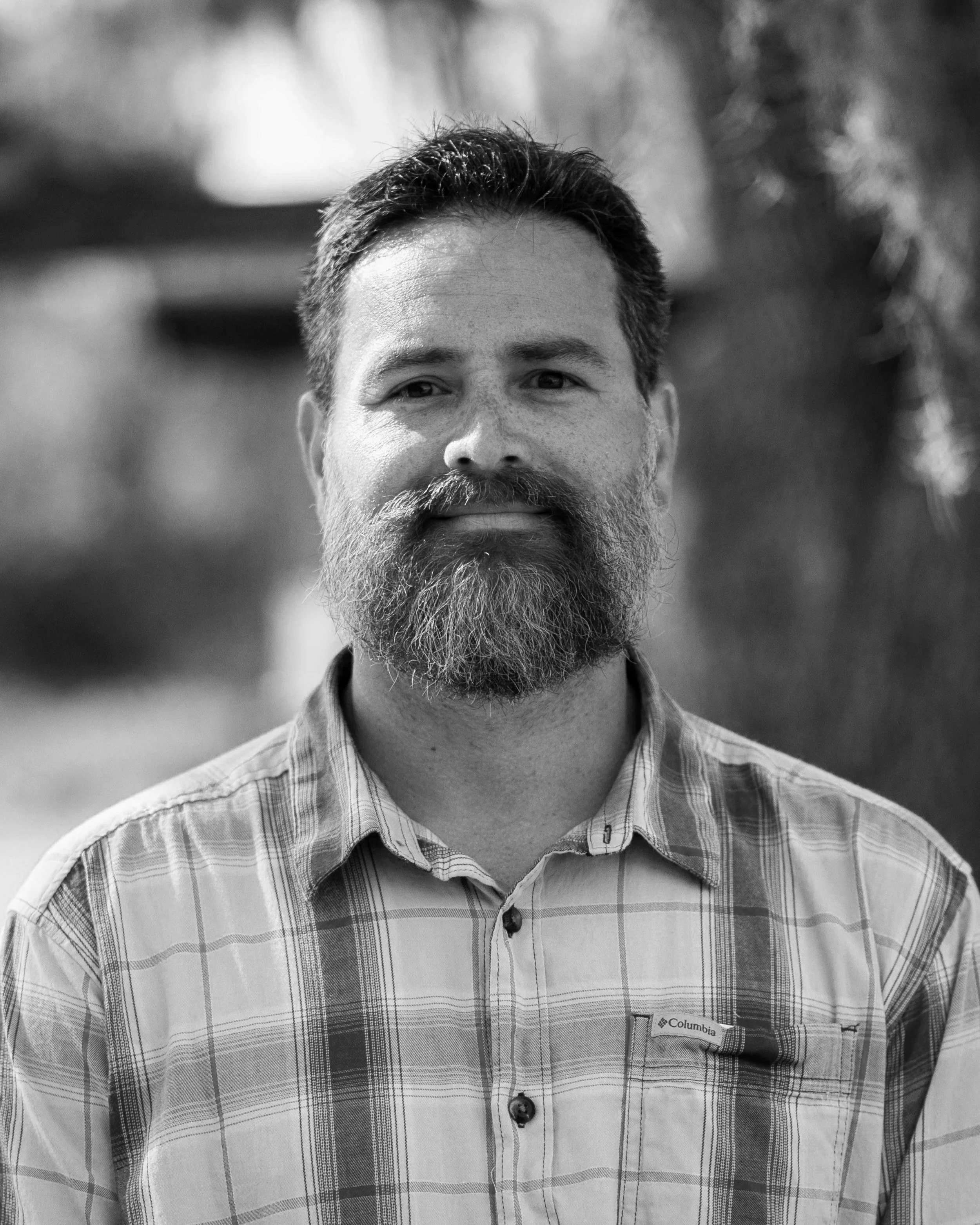 A man with a beard and mustache, wearing a plaid shirt, standing outdoors with blurred background.