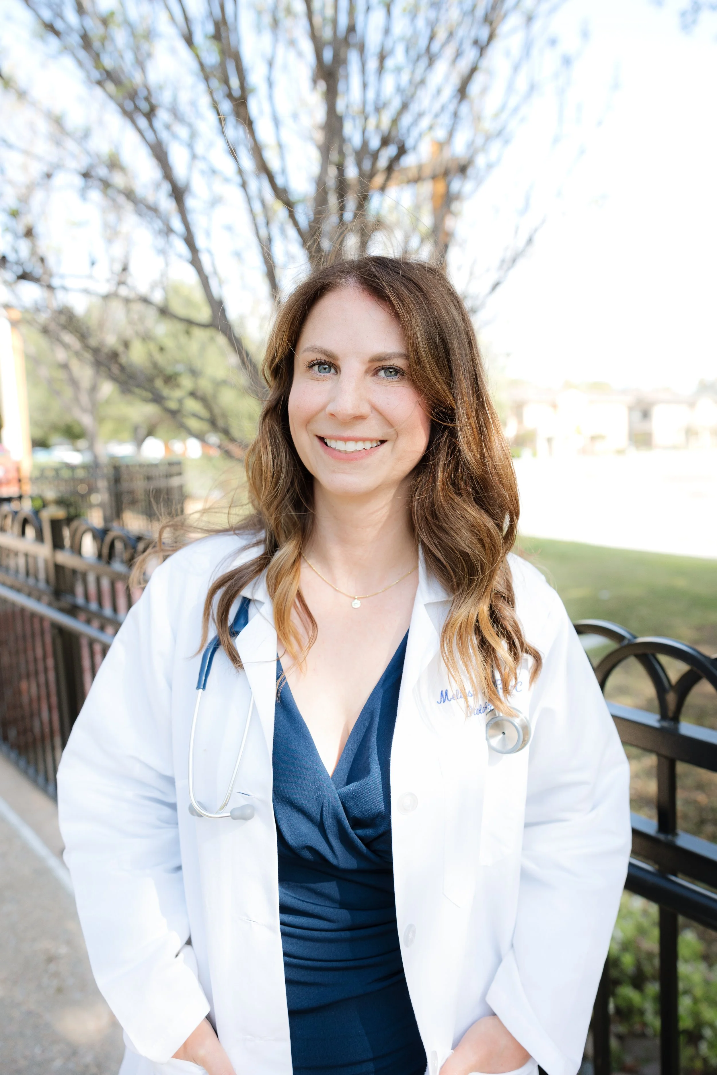 A young woman with long, wavy brown hair and blue eyes wearing a white medical coat and navy scrubs underneath, standing outdoors near a park bench with trees in the background. Melissa Spence, PA-C has been in clinical practice for 15 years.