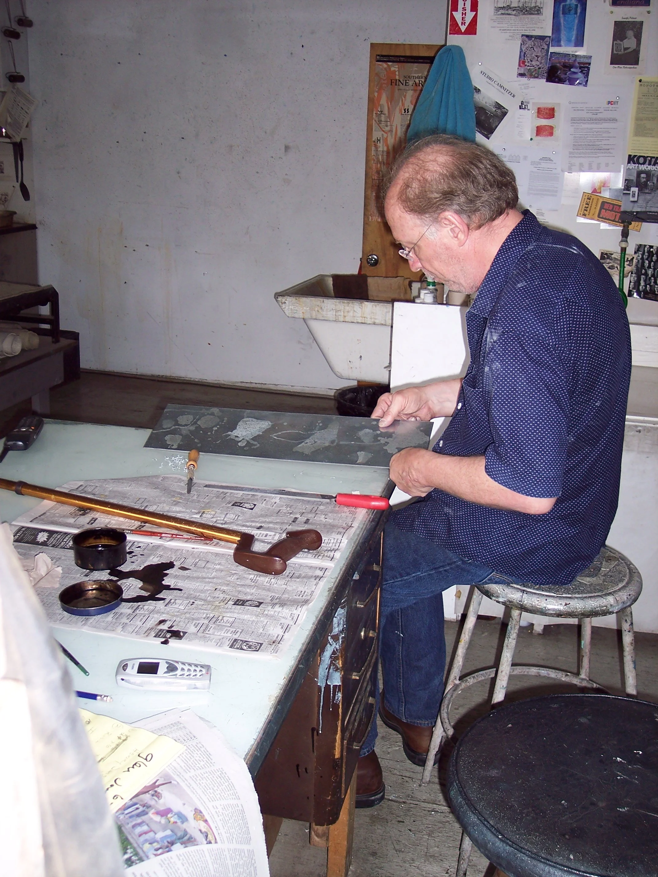 Glen Brunken at a cluttered table working on a craft project, surrounded by tools, newspapers, and papers in a workshop or studio setting.