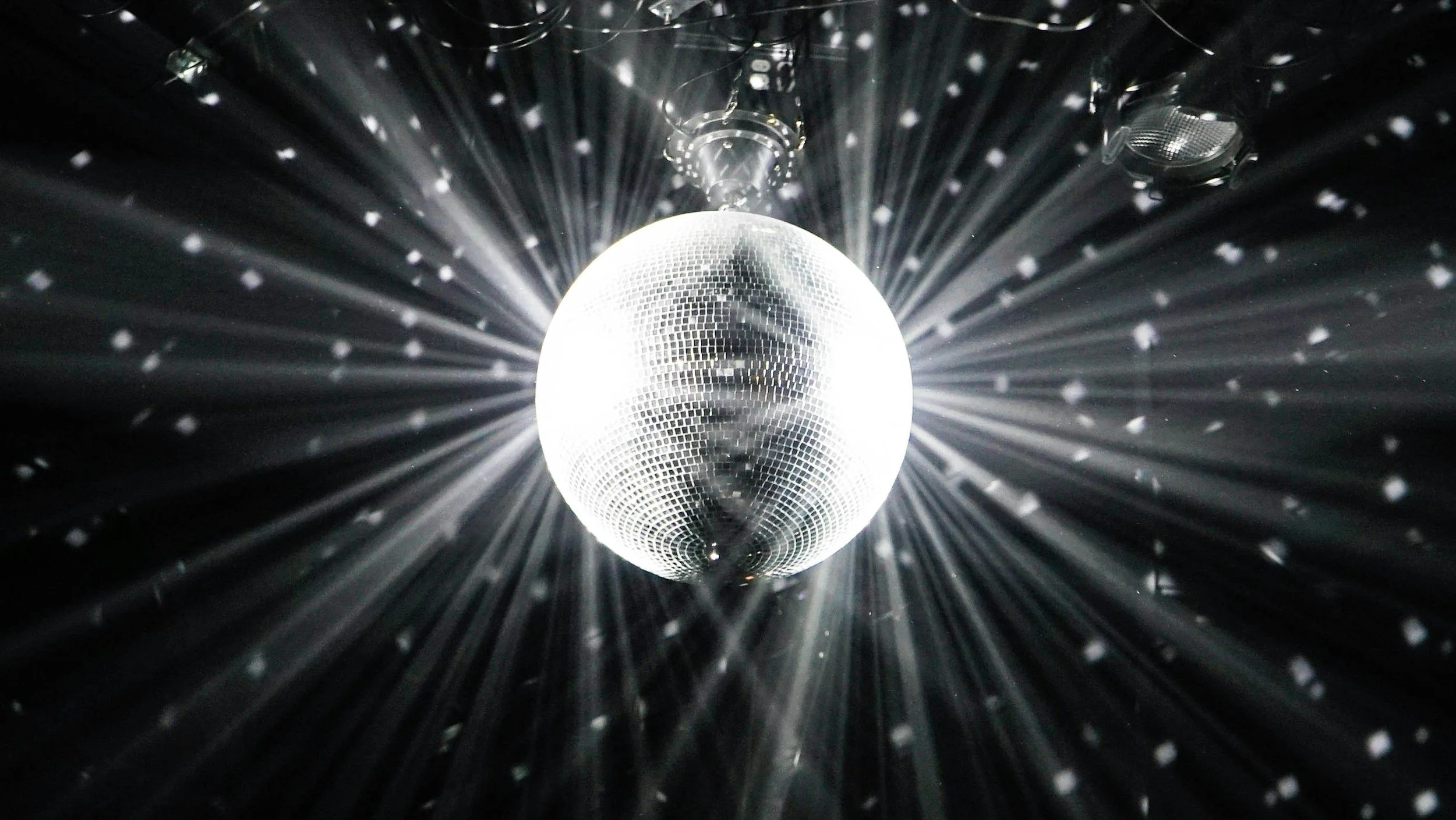 A person is reflected in a large disco ball hanging from the ceiling, with light beams radiating outward.