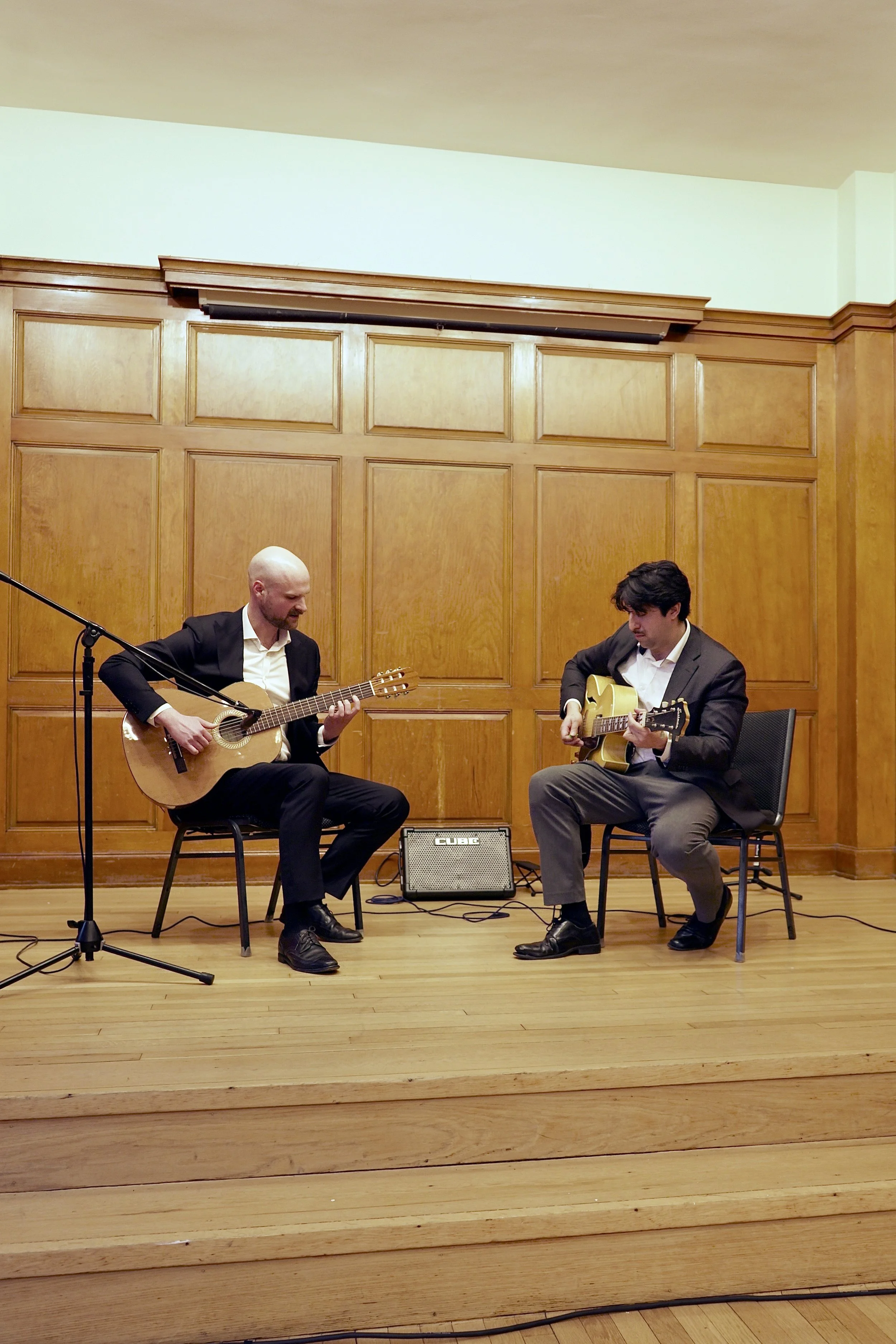 Two men in suits sitting on stage playing acoustic guitars, with a small guitar amplifier between them, in a wood-paneled room.