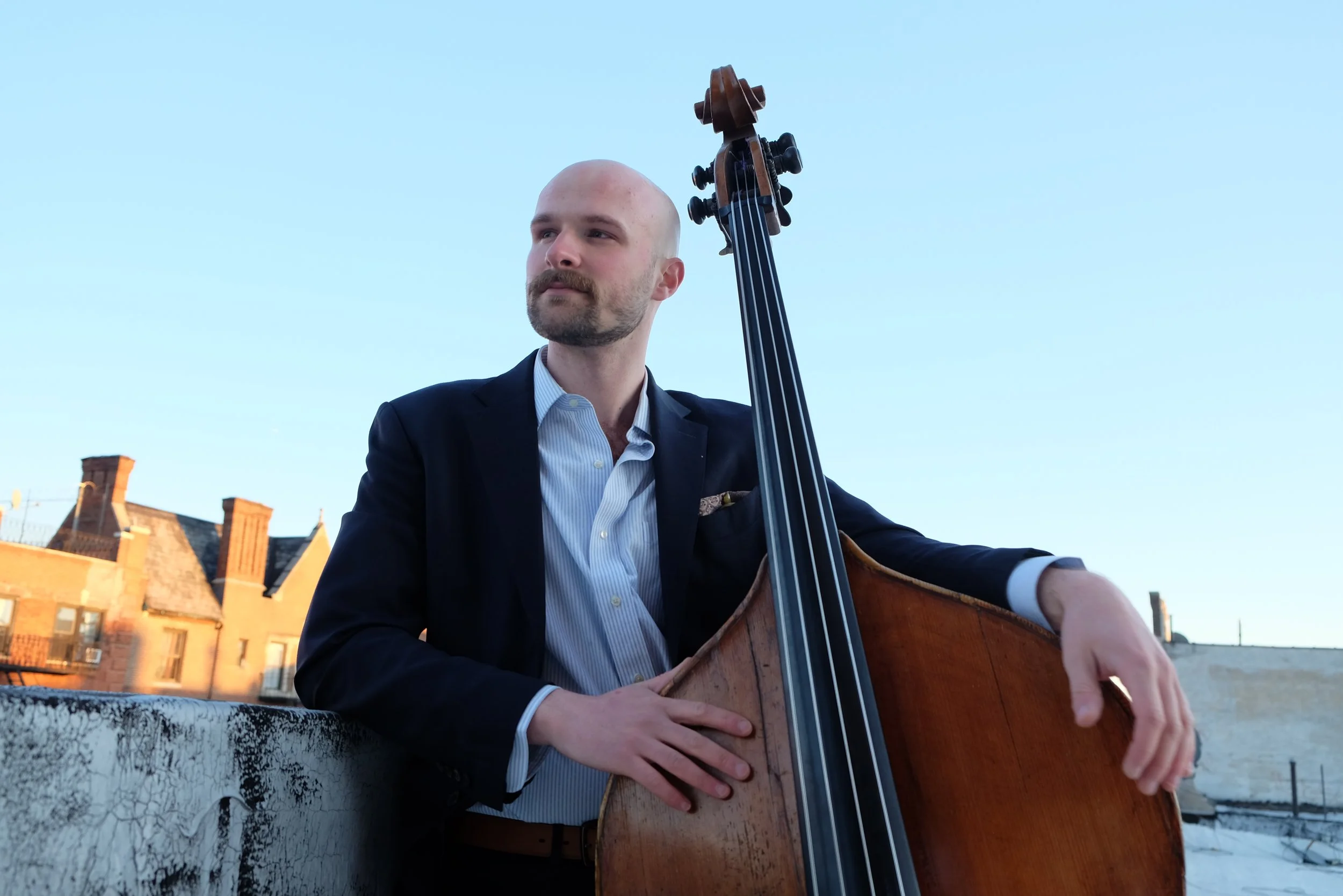 A man in a black suit jacket and light blue shirt playing a double bass outdoors against a clear blue sky, with brick buildings in the background.