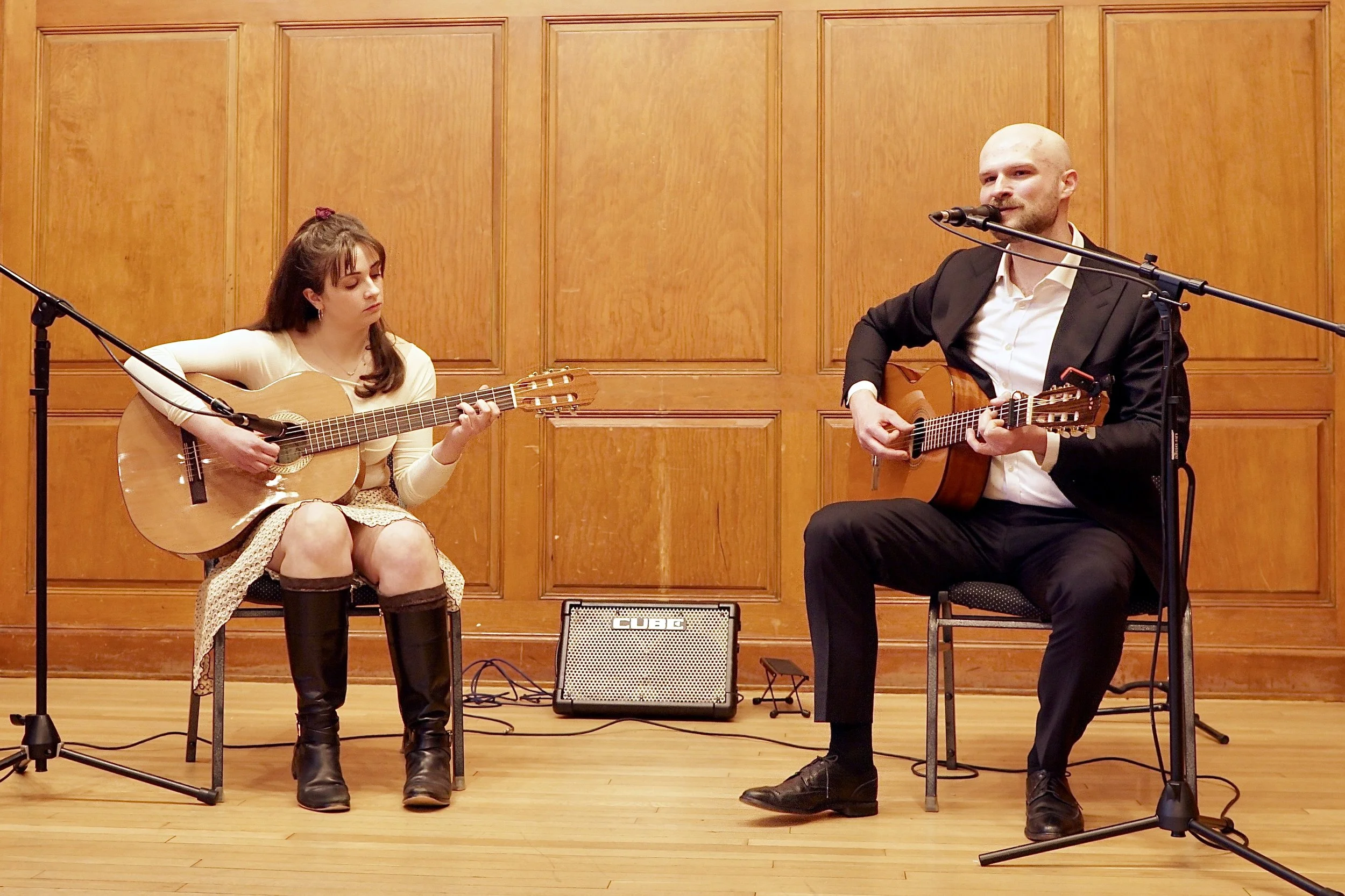 Young woman and man sitting on chairs playing acoustic guitars in a wooden-paneled room, with microphones and a small amplifier between them.