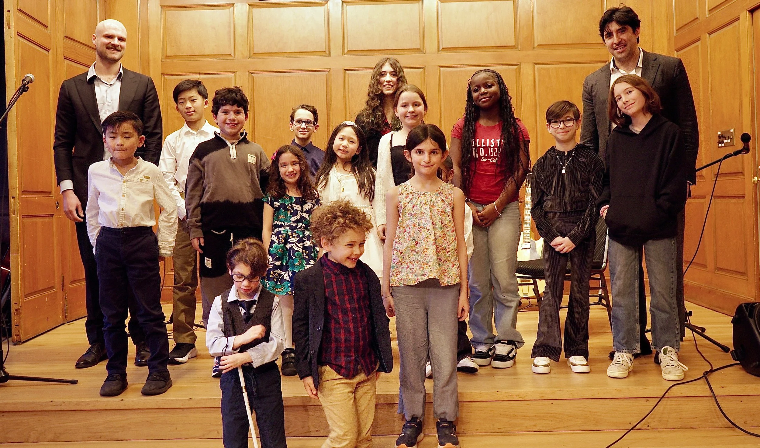 Group of children and two adult men standing on a wooden stage in a room with wood-paneled walls, smiling for a group photo.