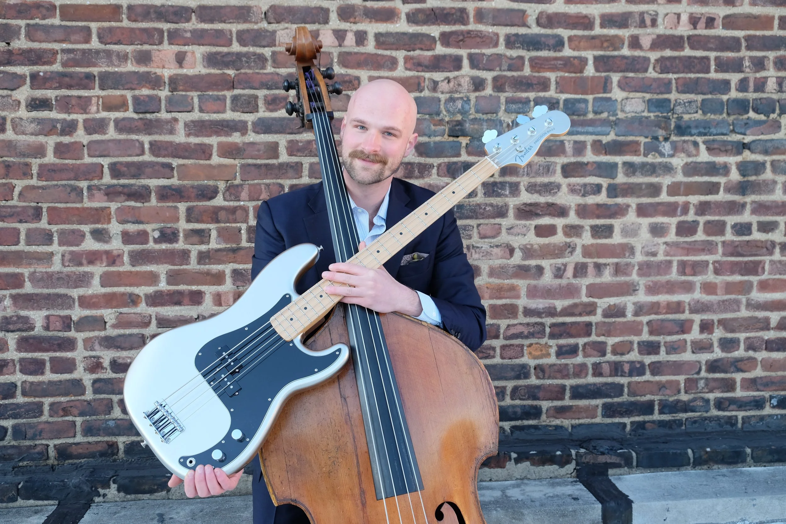 A man in a black suit holding a large double bass and a smaller electric bass guitar in front of a brick wall.