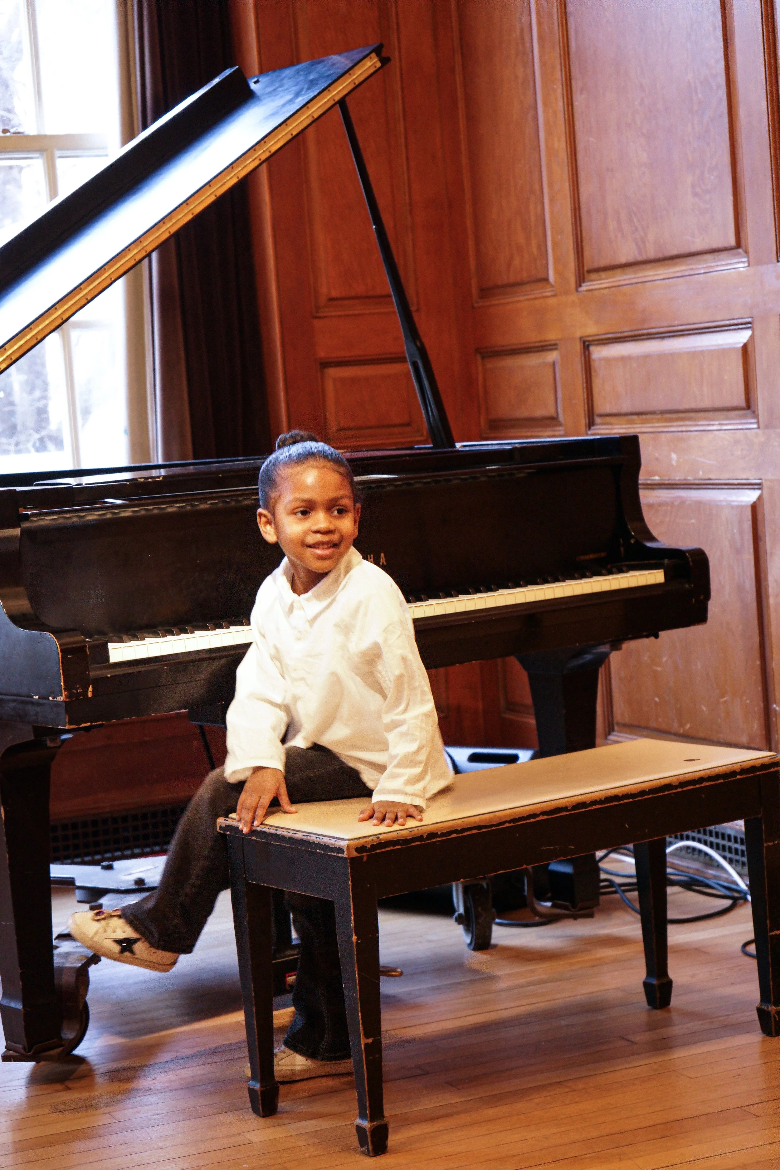 A young girl sitting on a bench in front of a grand piano in a wooden-paneled room.