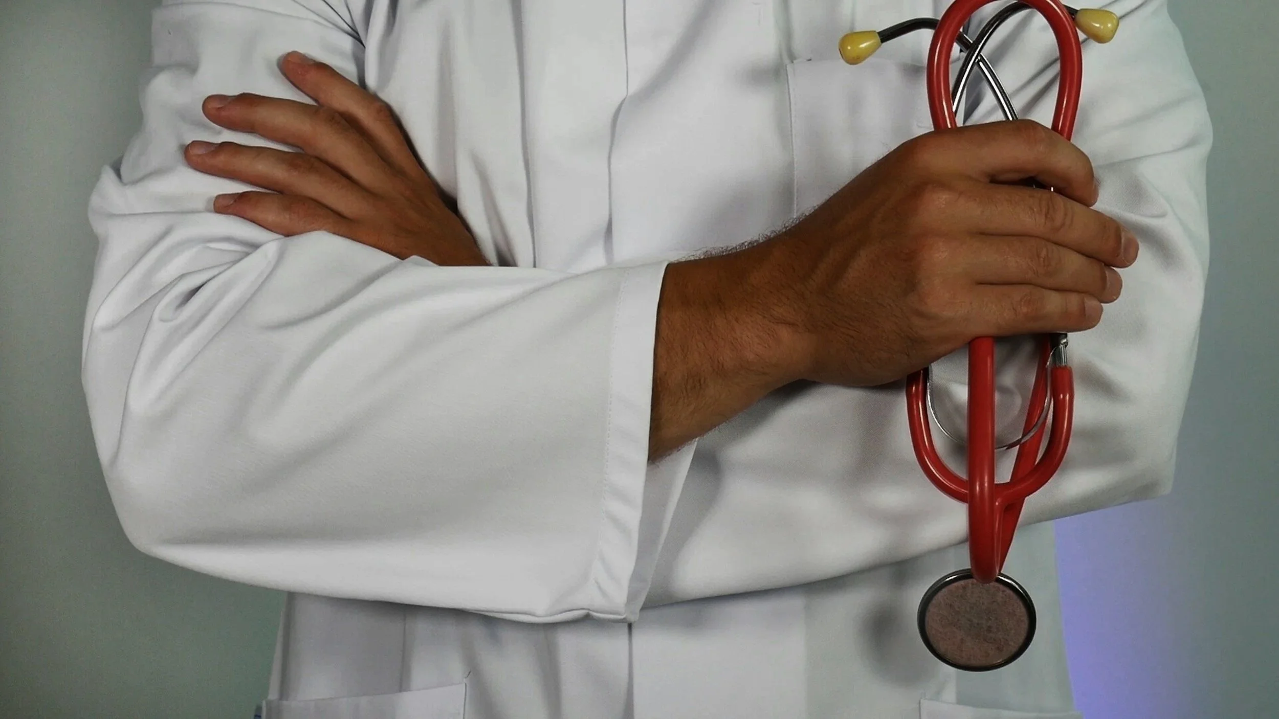 Close-up of a doctor or medical professional with crossed arms wearing a white coat, holding a red stethoscope.