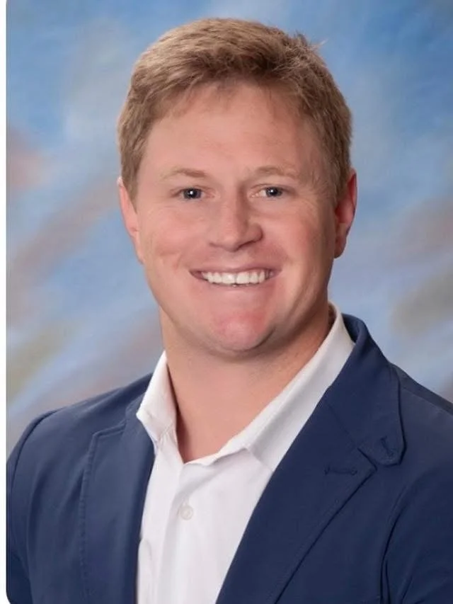 A portrait of a smiling man with light skin, short reddish hair, wearing a navy blue blazer over a white collared shirt, against a cloudy sky background.