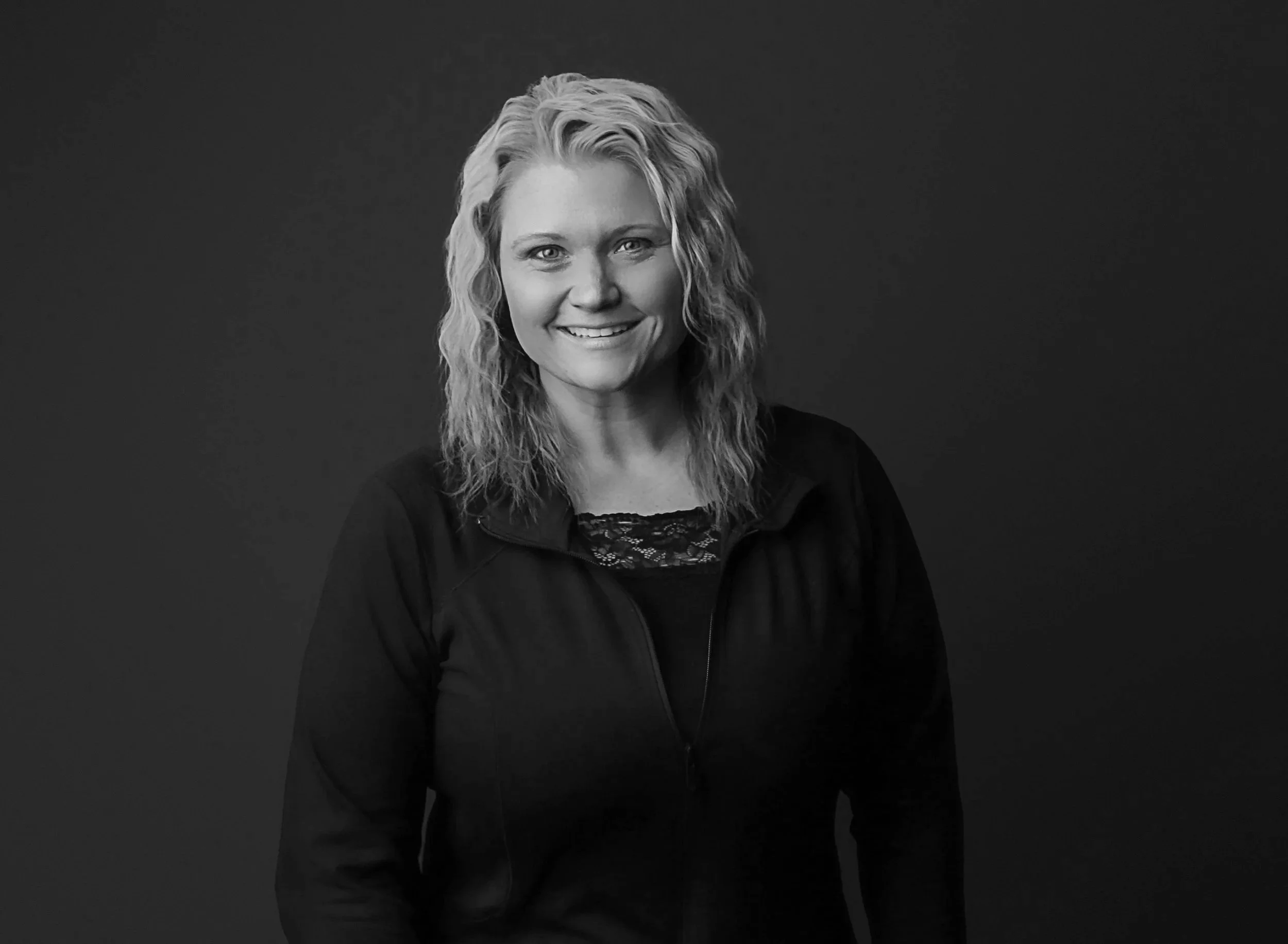 A woman with blonde, wavy hair smiling, wearing a black jacket and lace top, against a plain dark background.