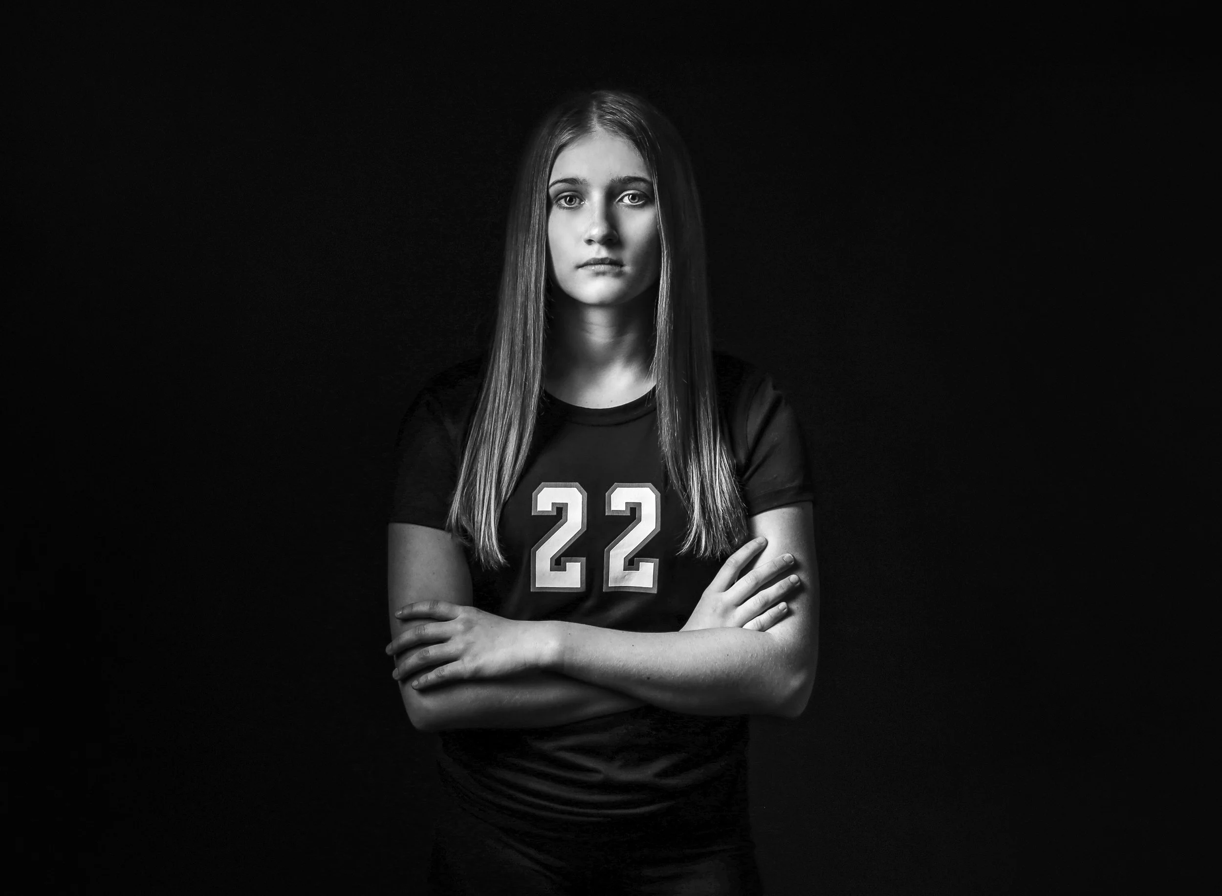 Black and white photograph of a young woman with long straight hair, crossing her arms, wearing a sports jersey with the number 22, standing against a black background.