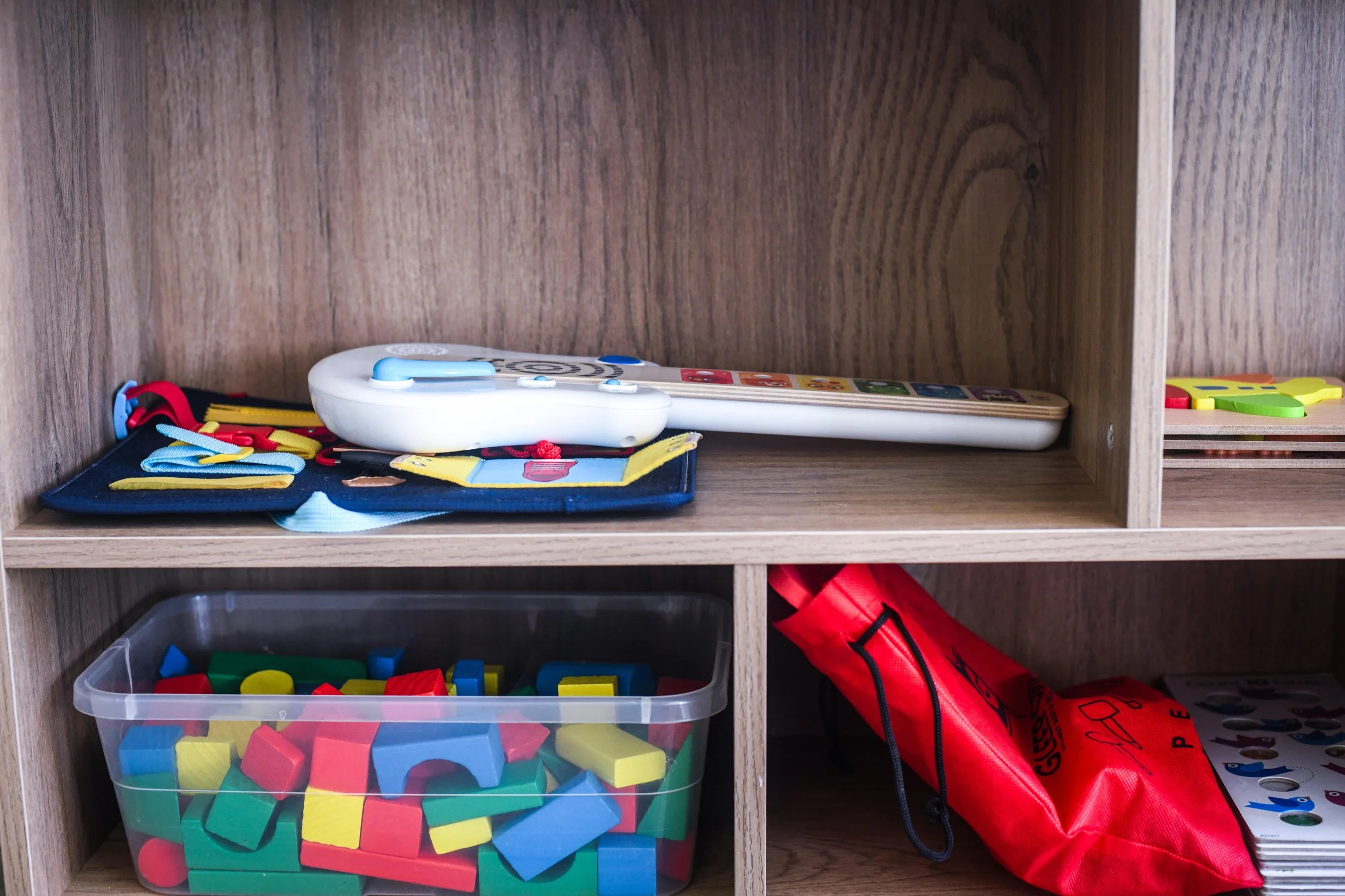 A wooden shelf containing toys and educational materials, including a container of colorful foam blocks, a toy phone, a red and black bag, and various other toys and learning tools.