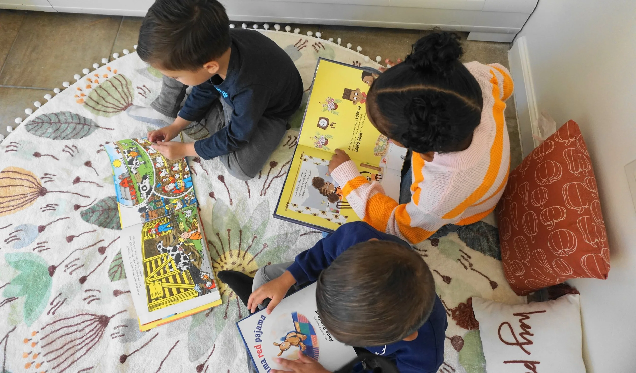 Three children sitting on a colorful rug, reading children's books. The books contain illustrations of animals, objects, and characters for early learners.