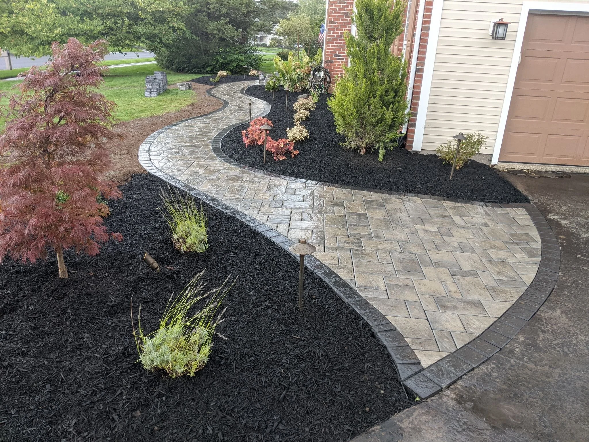 A winding stone walkway leading away from a house, surrounded by freshly mulched flower beds with small plants and bushes.