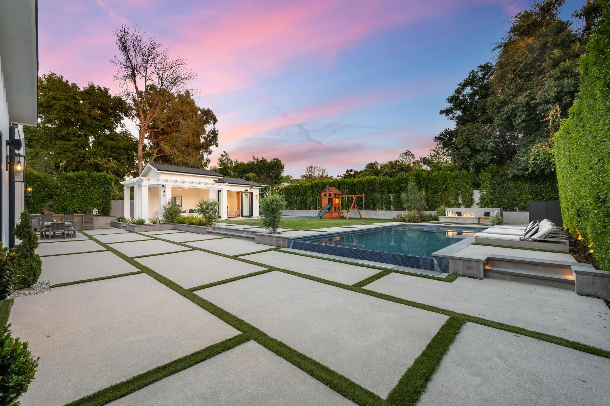 Modern backyard with a geometric concrete patio, green grass inlays, a swimming pool, outdoor furniture, a children's play structure, and a sky at sunset.