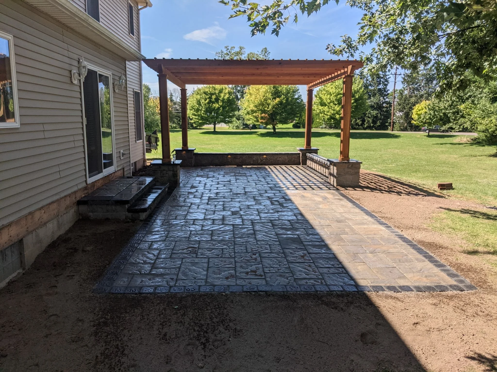 Backyard patio with newly paved stone surface, wooden pergola framing a view of green lawn and trees, part of house with beige siding, sliding glass door, outdoor lighting fixtures, and a small wooden seating bench.