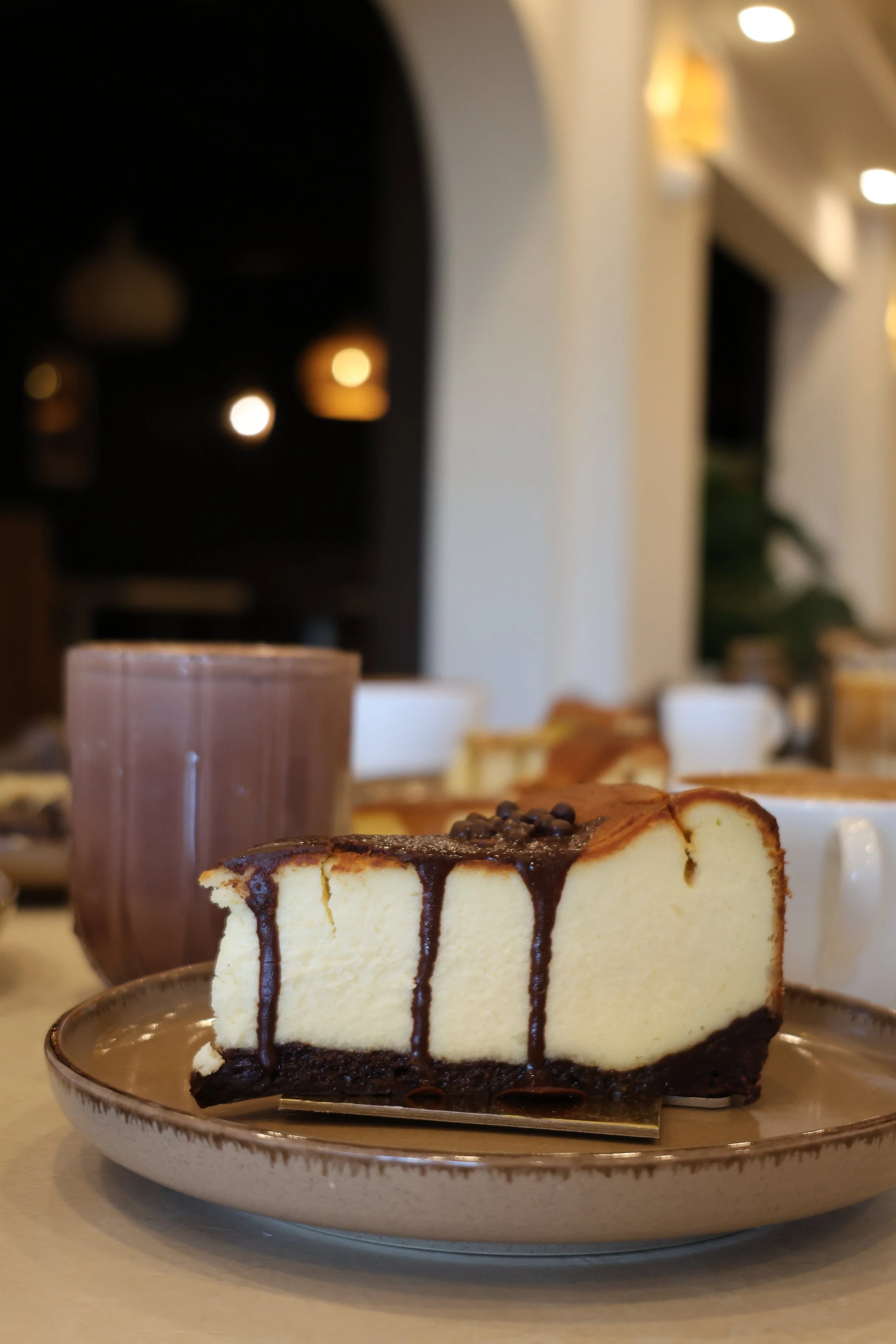 A slice of cheesecake with chocolate topping and drizzle, served on a beige plate. A glass of chocolate dessert and other blurred desserts and a cozy indoor setting are in the background.