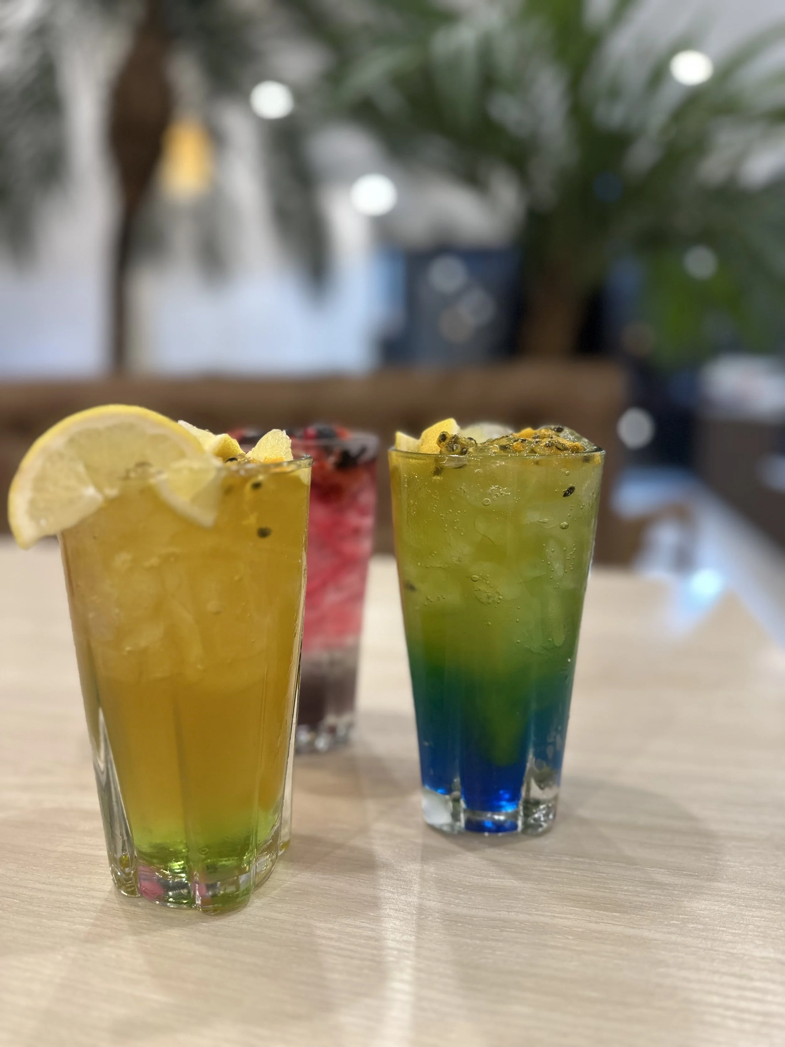 Three colourful layered mocktails in tall glasses with lemon garnish on a light wooden table.