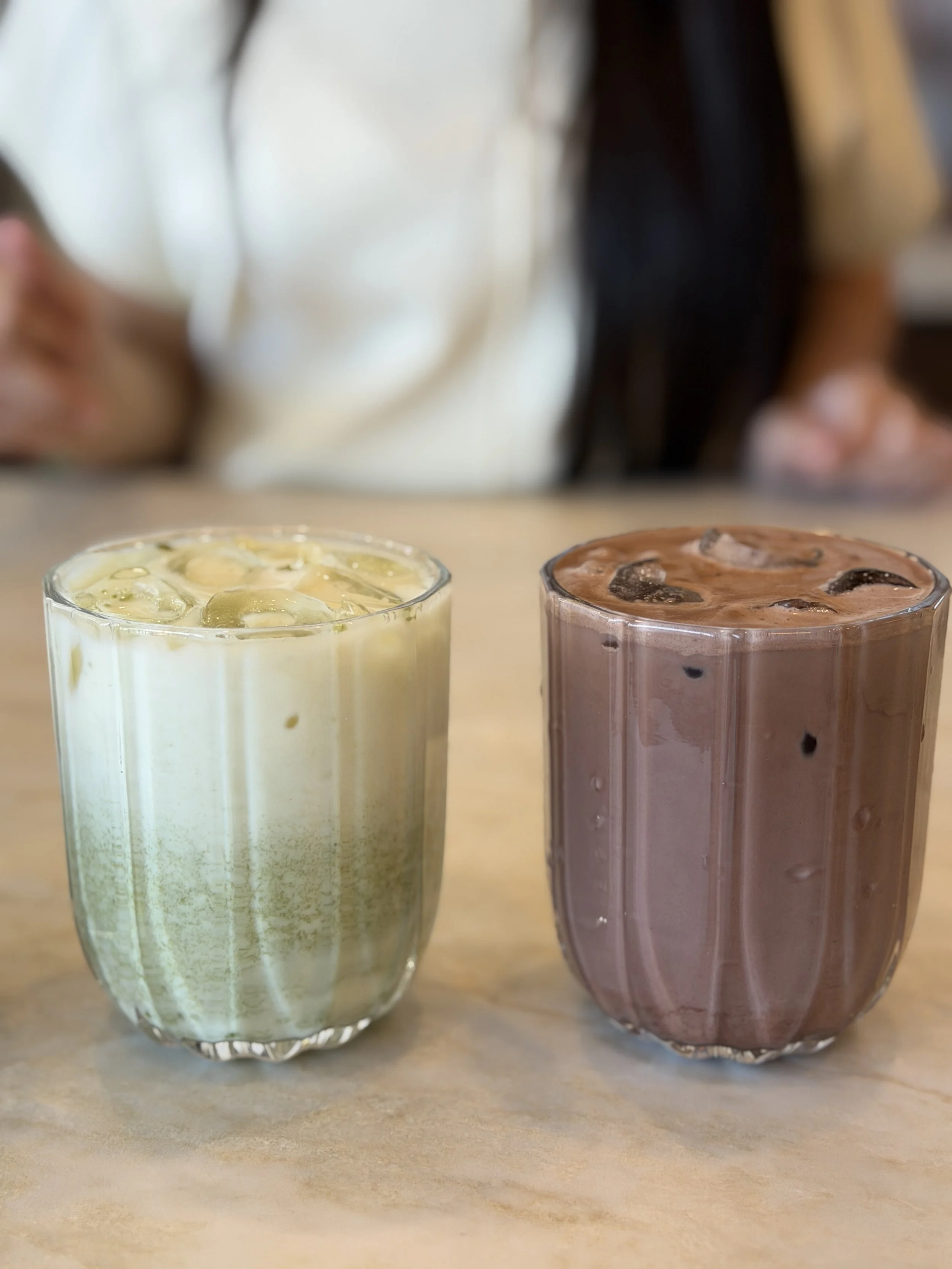 Two glasses of iced beverages on a wooden surface, one with a creamy color and the other with a chocolate color, both with ice cubes.