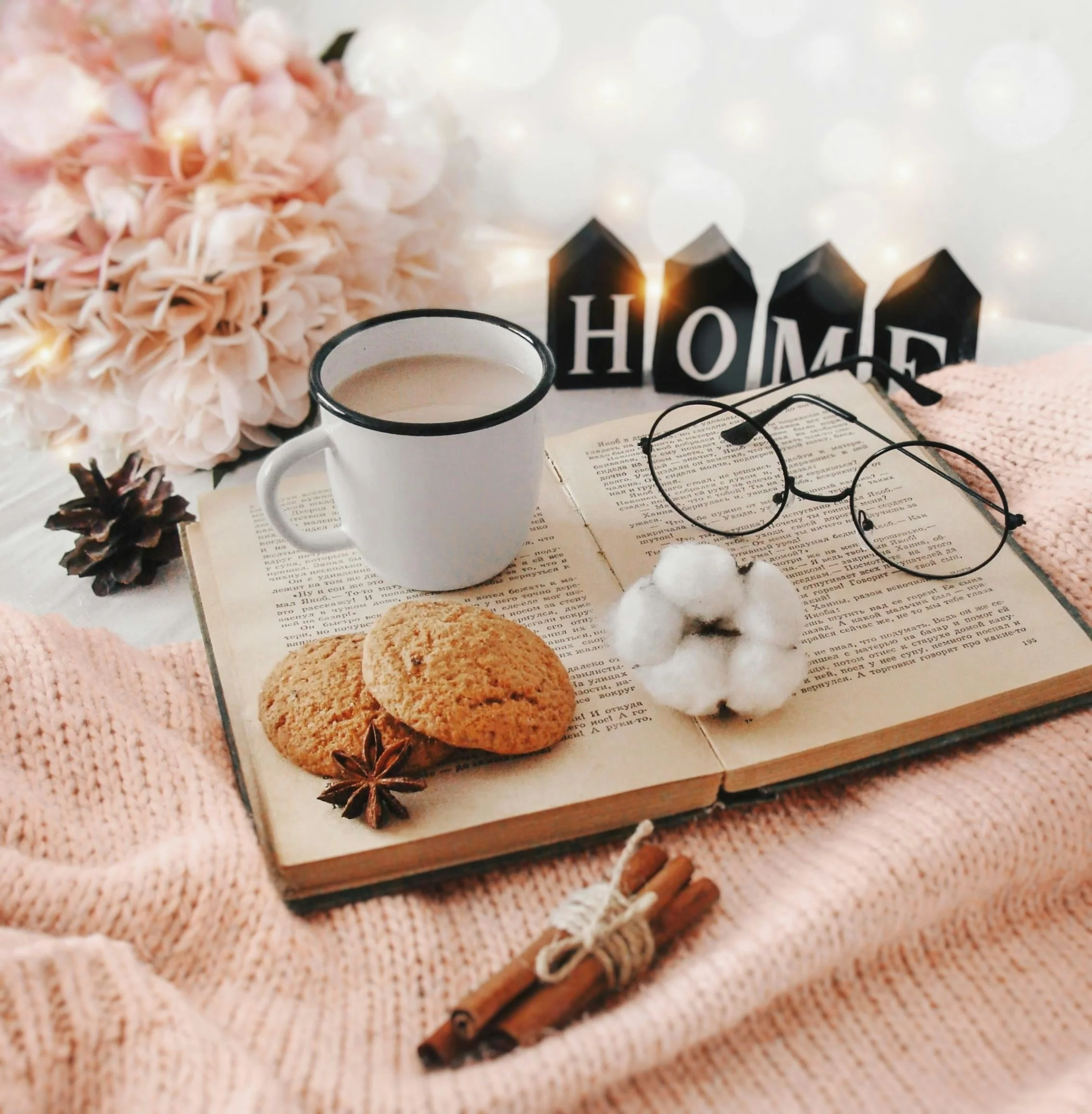A cozy scene with a pink flower, a white mug with a hot beverage, eyeglasses, an open book, and small decorative items including pine cones, star anise, and a wooden sign spelling 'HOME' in black houses, all arranged on a soft pink knitted blanket.