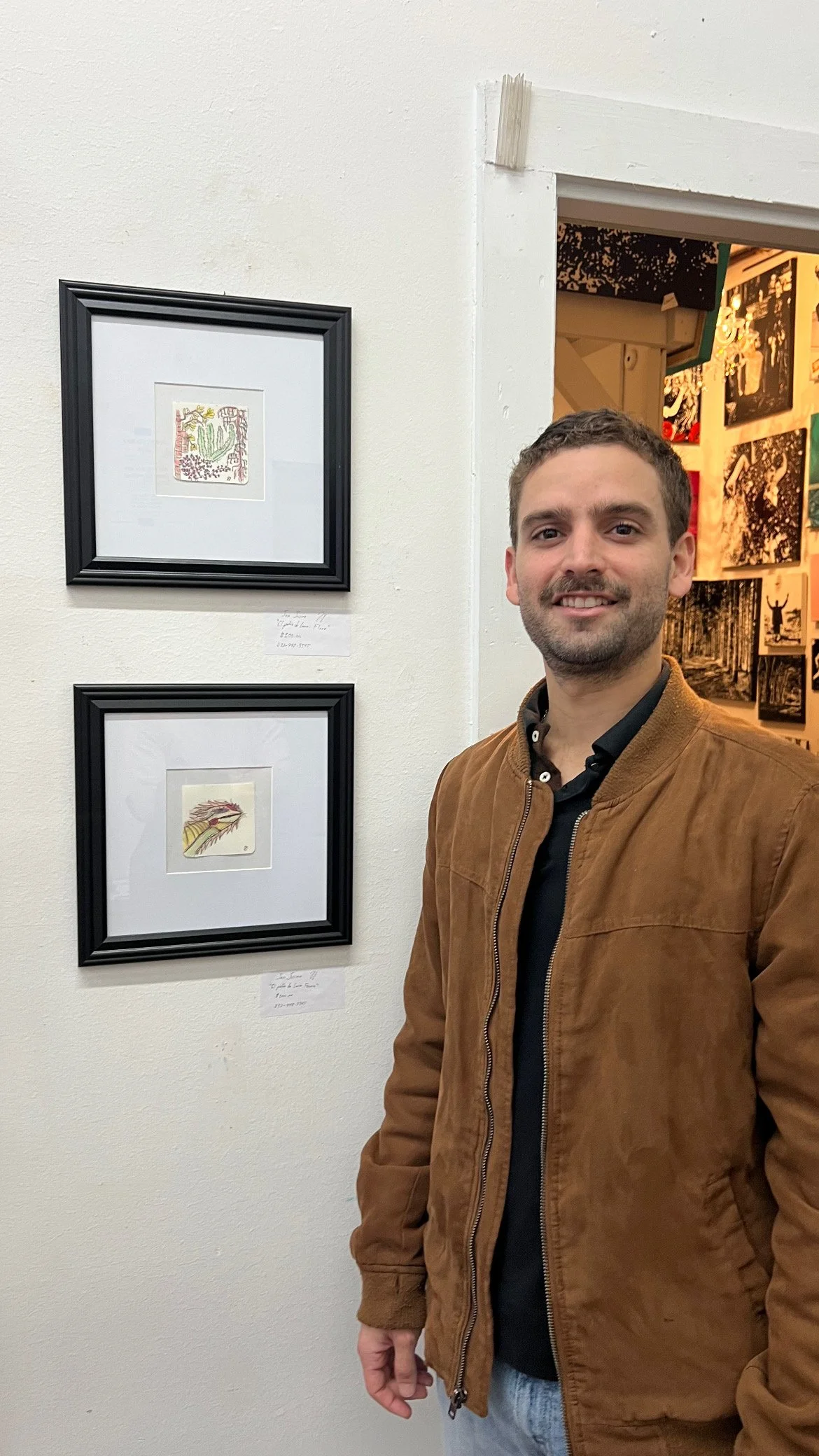 A man with a mustache and beard smiling at the camera in an art gallery, standing beside two framed pieces of artwork on a white wall.