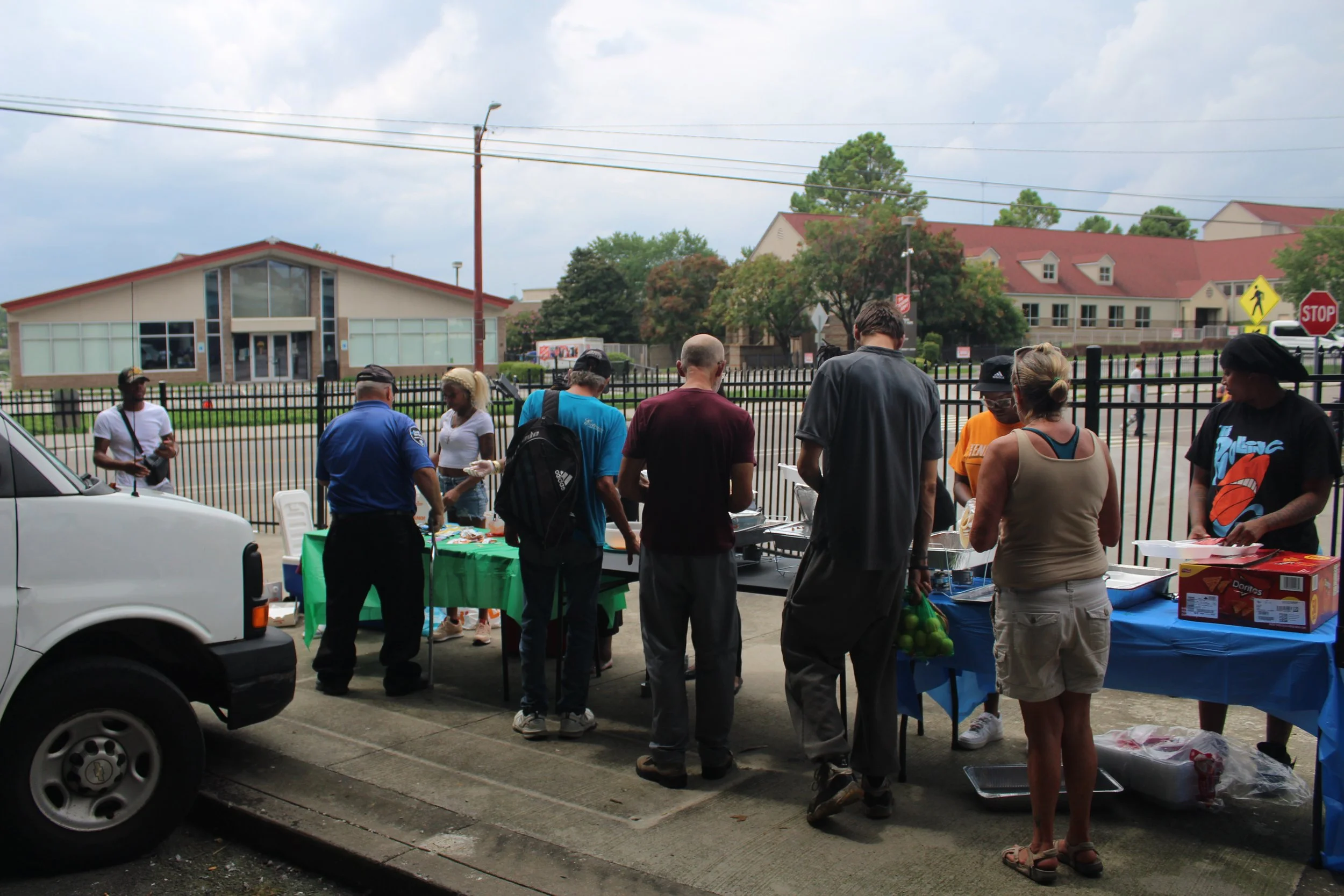 People gathered around tables with food and drinks outside behind a black fence, with a building and trees in the background.