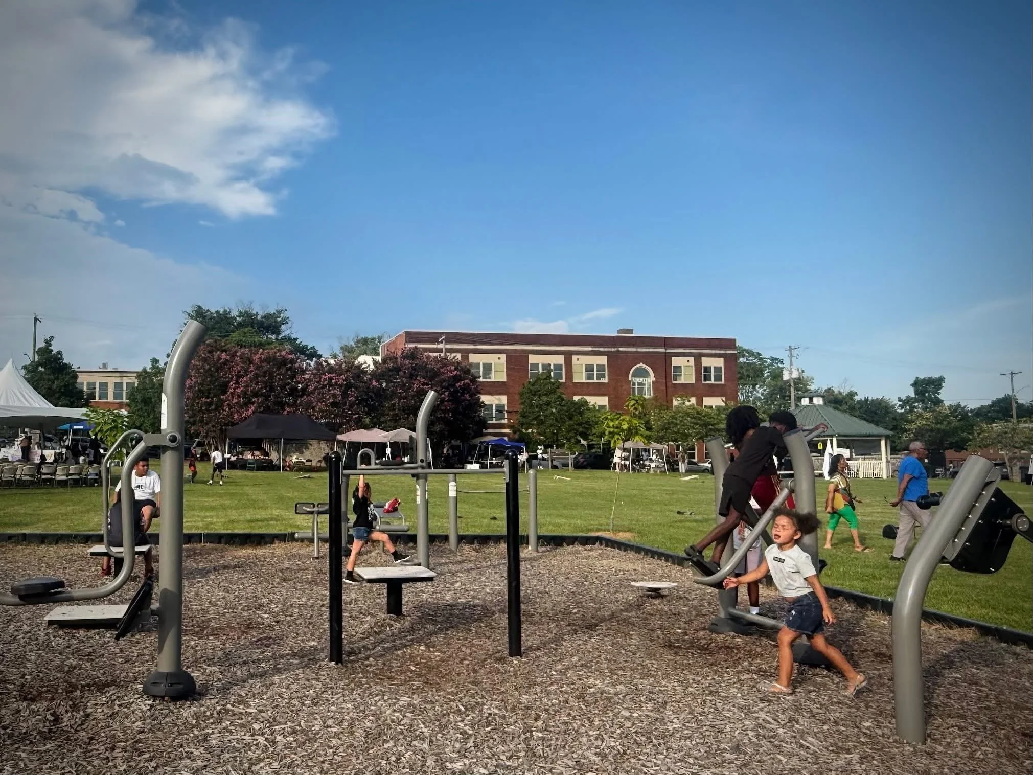 Children playing on outdoor exercise equipment in a park with tents, trees, and a multi-story brick building in the background.