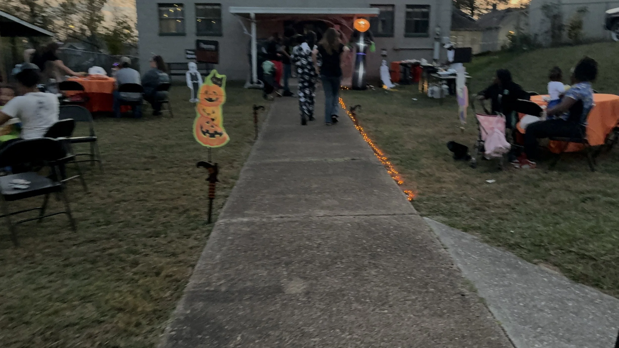 A Halloween-themed outdoor party at dusk with tables covered in orange tablecloths, Halloween decorations, and people gathered around in front of a house. There are Halloween signs and string lights along the sidewalk.