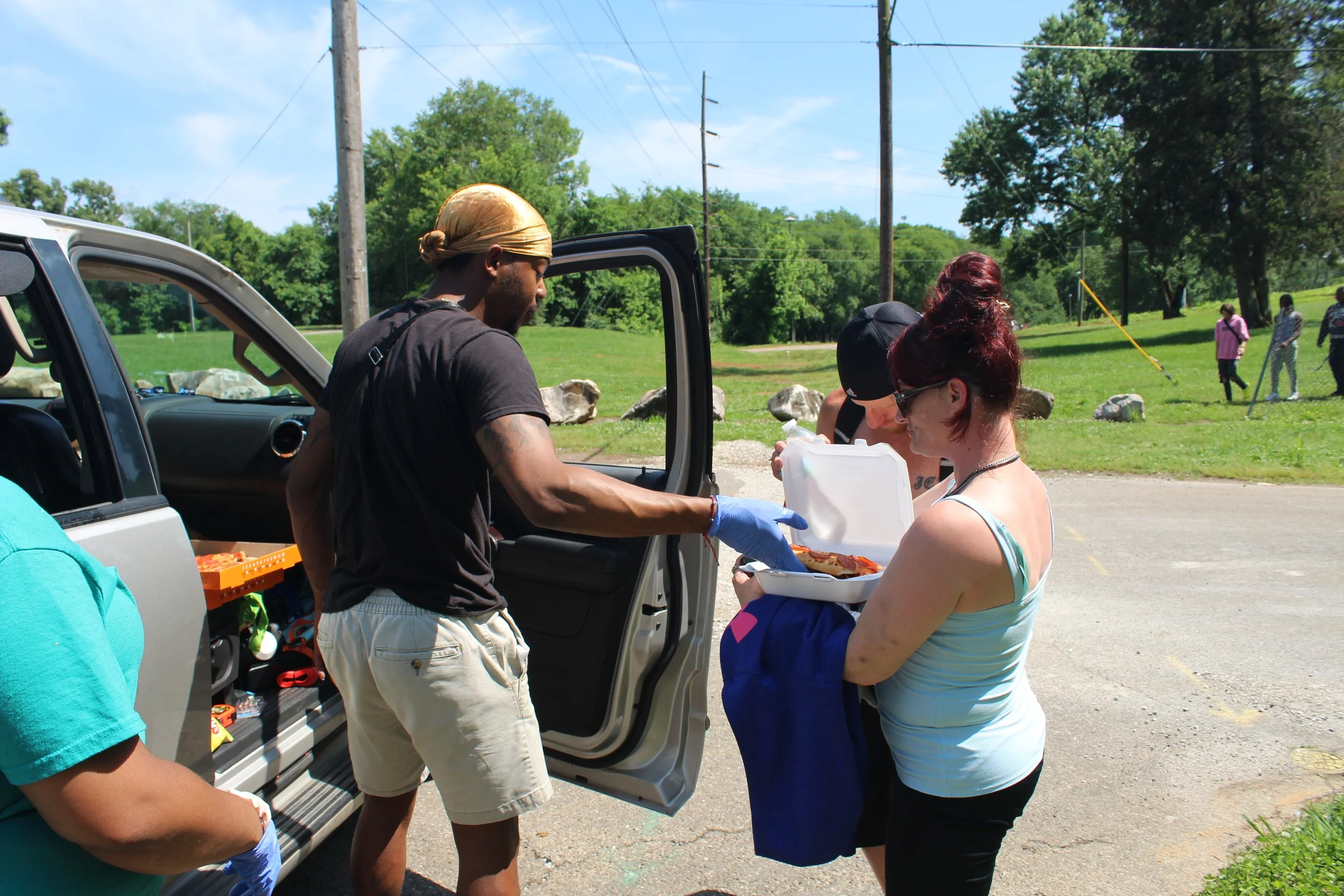 People receiving food from a person standing outside a vehicle in a park-like area.