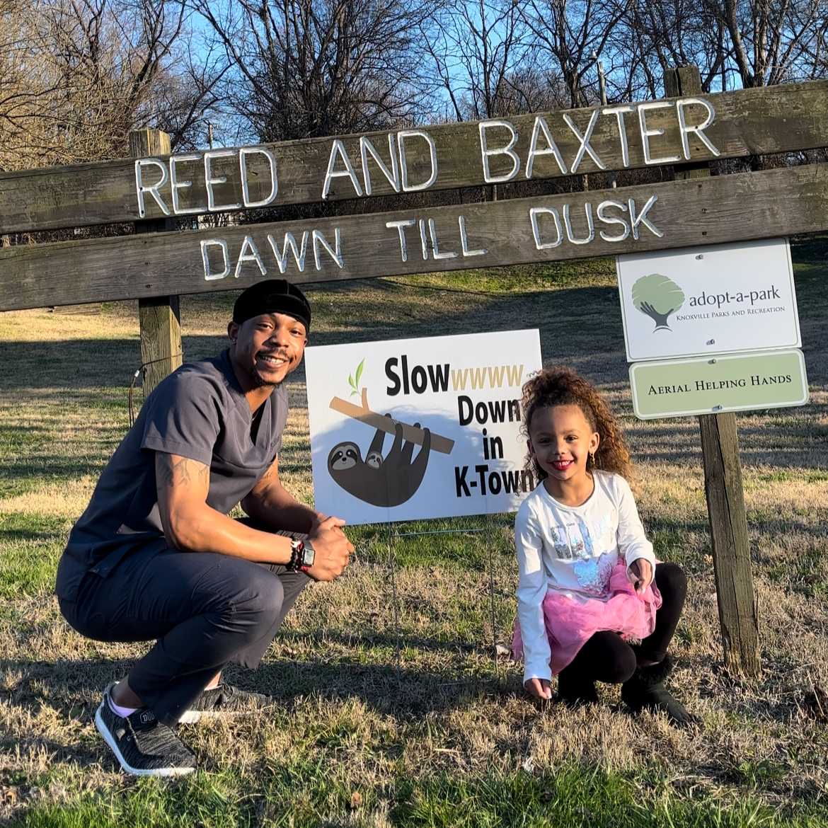 A man and a young girl smiling and crouching by a park sign that reads "Reed and Baxter Dawn till Dusk." There is a sign with a sloth hanging from a tree branch, and a smaller sign indicating "adopt-a-park" and "Aerial Helping Hands." The scene is ou