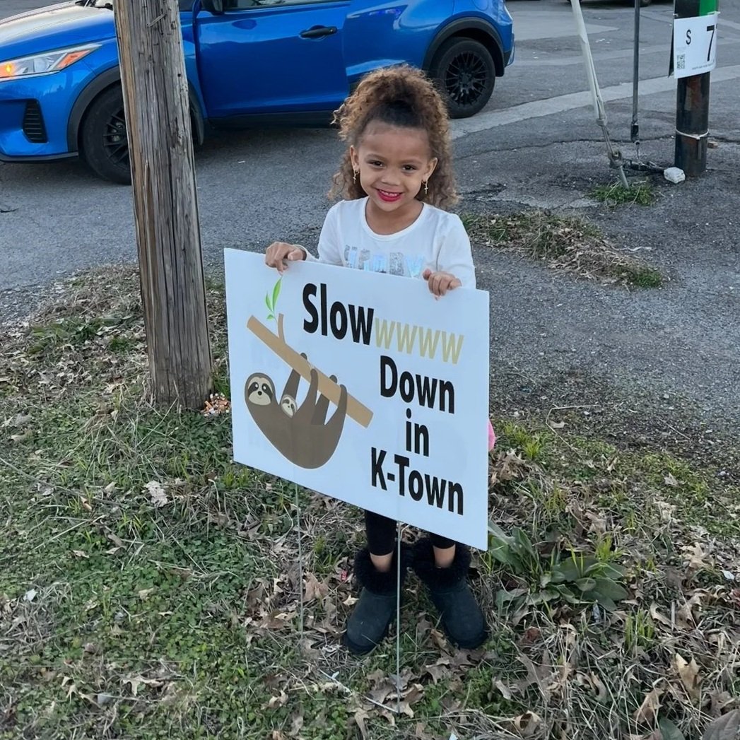 A young girl with curly hair holding a sign that says "Slow WWWW Down in K-Town" with a picture of a slow-moving sloth hanging from a tree branch. She is smiling and standing outdoors near a wooden pole, with cars visible in the background.