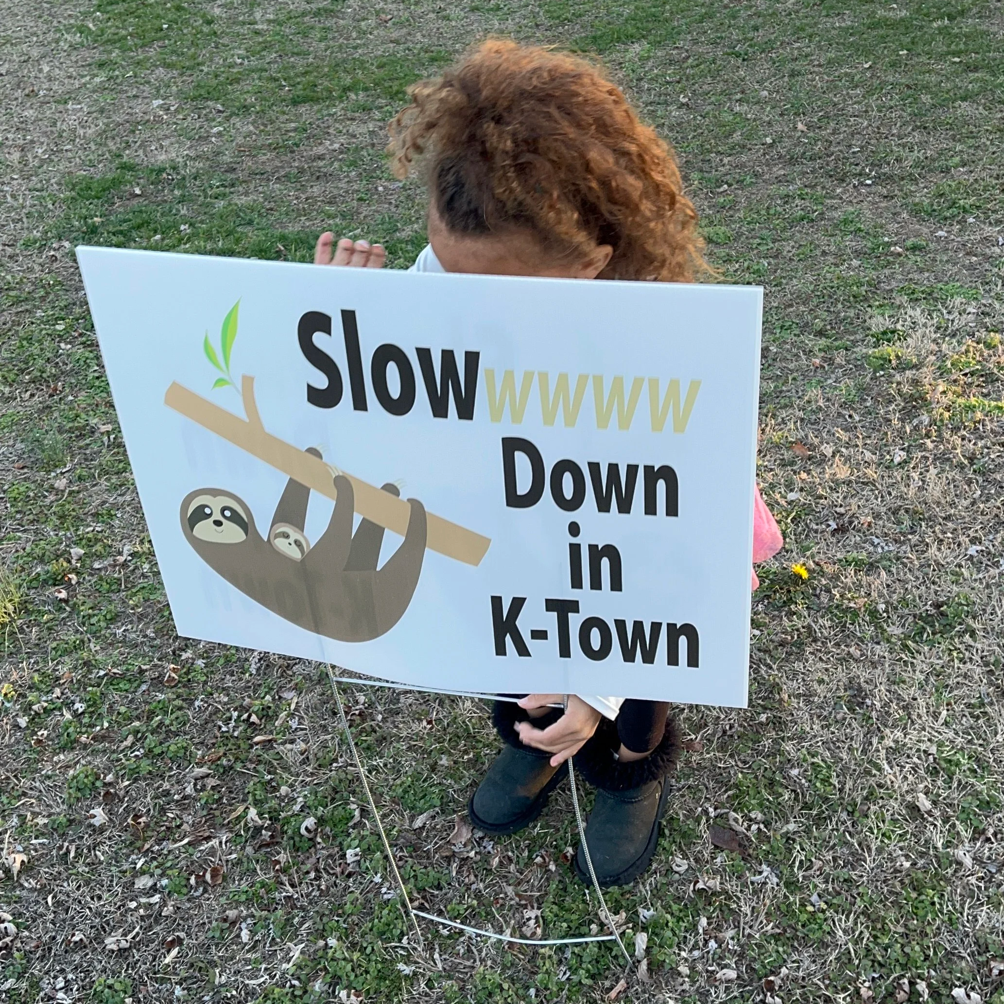 A child holding a sign that reads 'Slow WWW Down in K-Town' with an illustration of two sloths hanging from a tree branch.