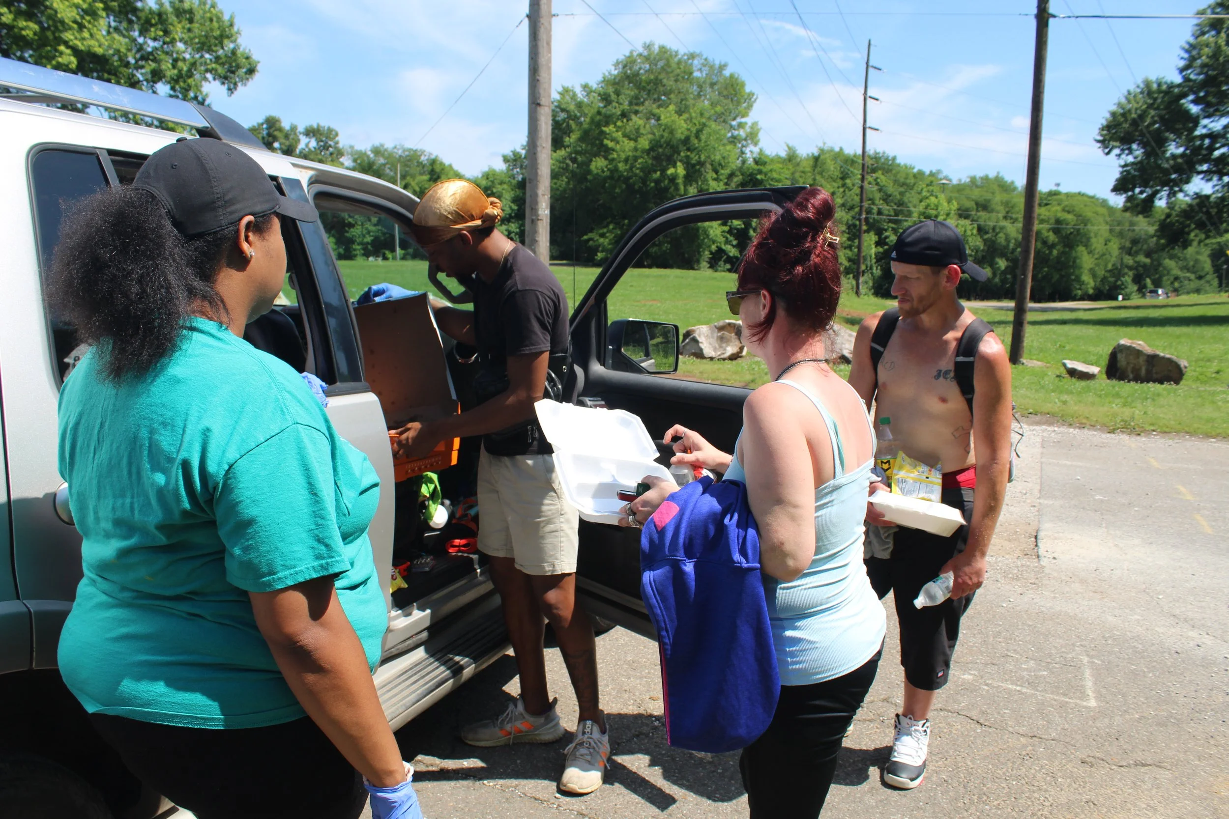 Group of four people buying food from a street vendor in a parking lot on a sunny day, with green trees and power lines in the background.