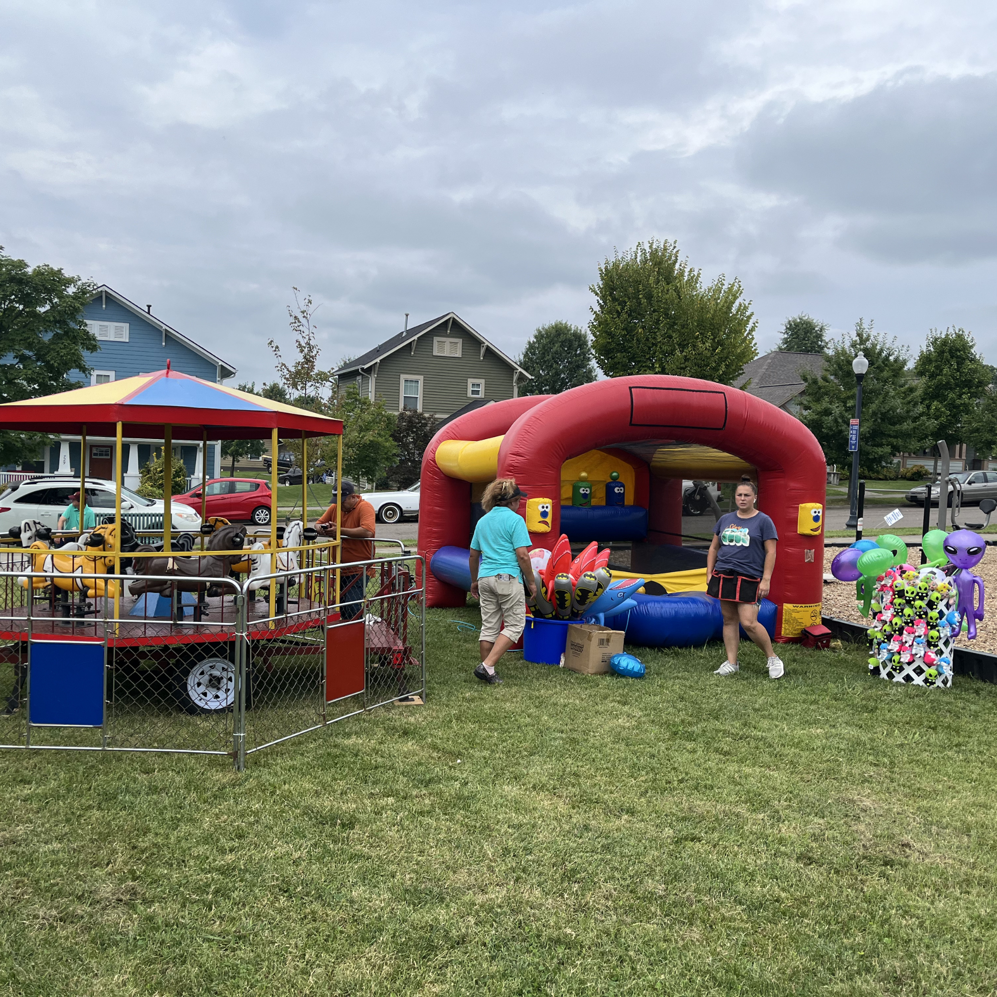 Kids and adults at an outdoor carnival with inflatable bounce house, carousel rides, and colorful decorations on a cloudy day.