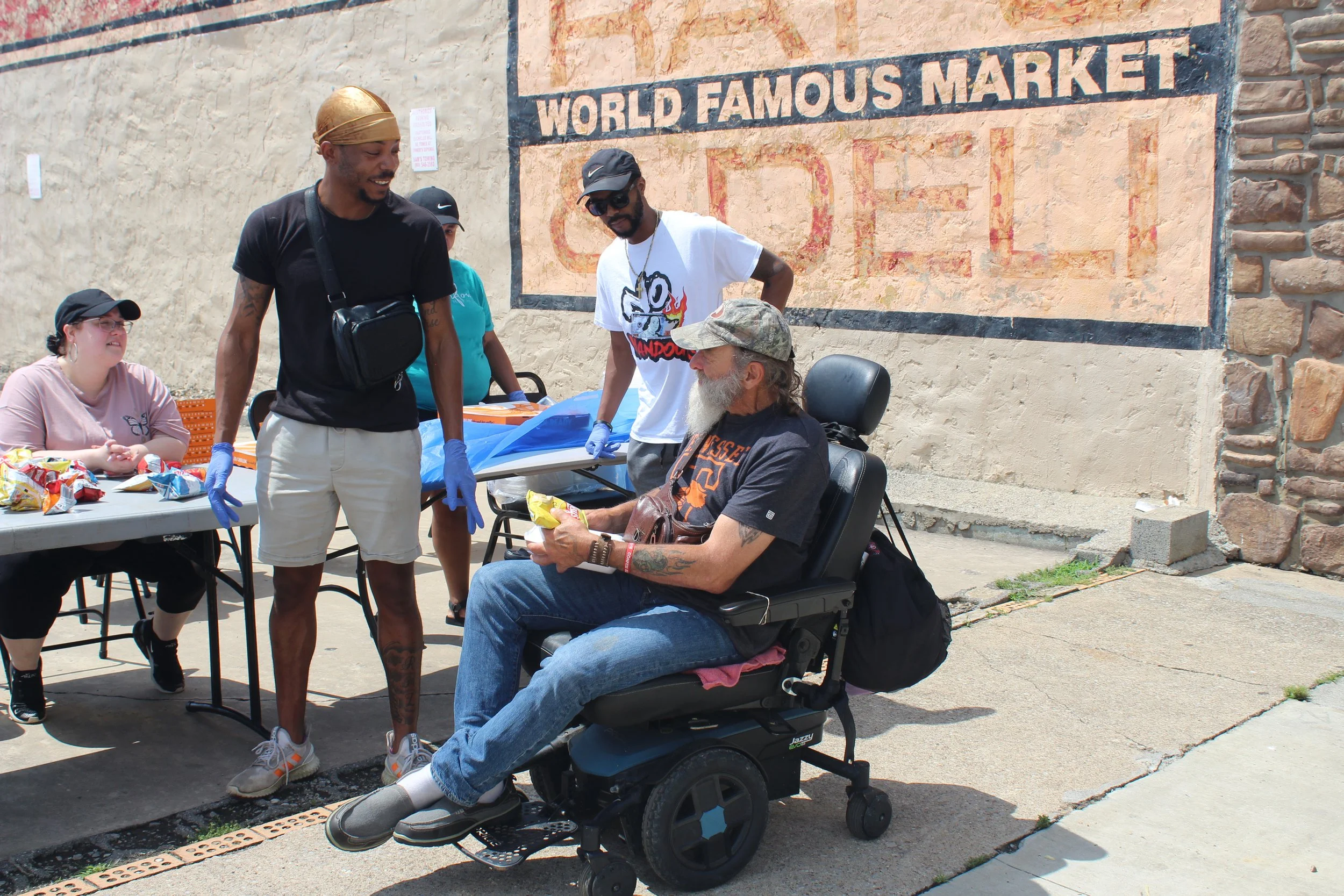 A man with a beard and sunglasses sitting in a power wheelchair, receiving a snack from a smiling man in a black t-shirt and beige shorts. Two women are sitting at a table with snacks nearby, and two other men are standing nearby, outdoors near a wal