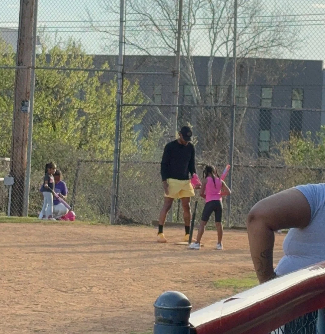 A man coaching a young girl in baseball on a dirt field, with two girls playing in the background near a chain-link fence and trees.
