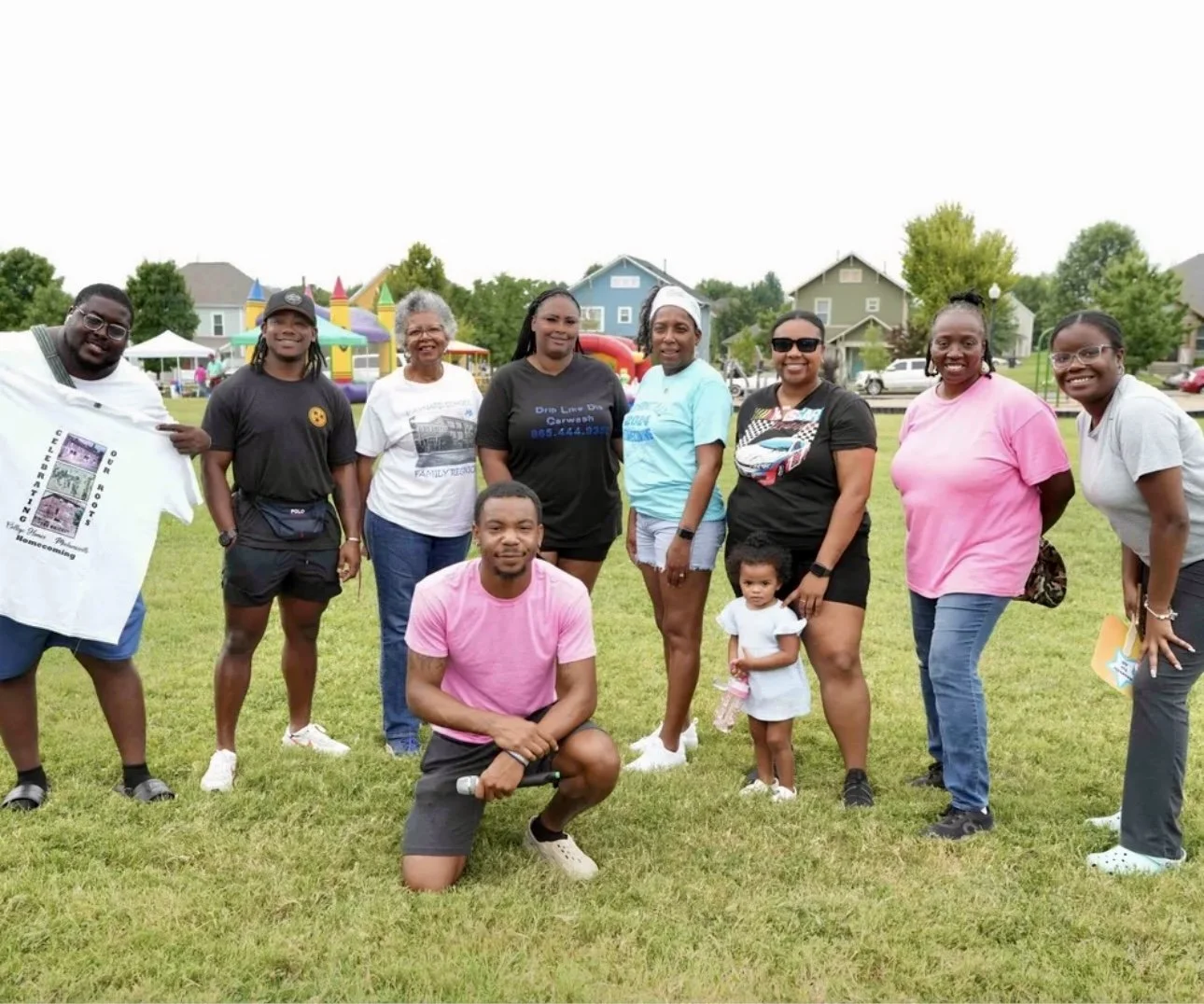 Group of ten people and a child posing on a grassy field at an outdoor event with play structures and tents in the background.