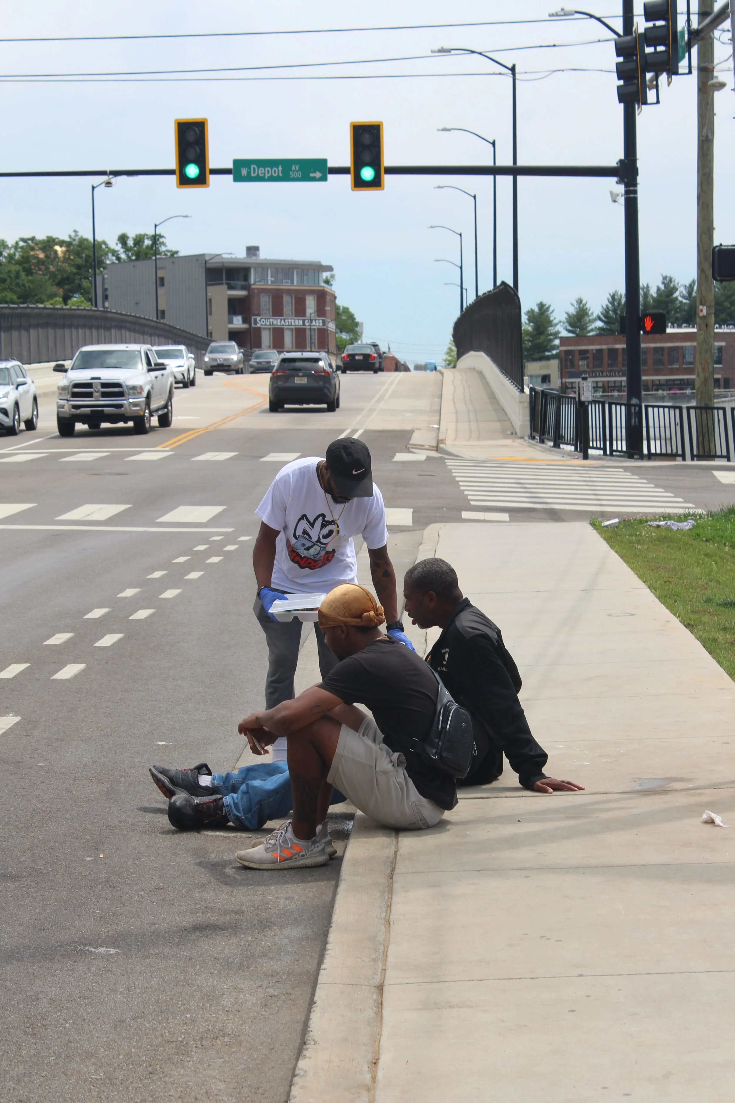 Three men sitting and standing on a sidewalk at a street intersection, with traffic lights and cars in the background. One man is sitting, another is standing with papers in his hands, and a third man is sitting on the edge of the sidewalk.