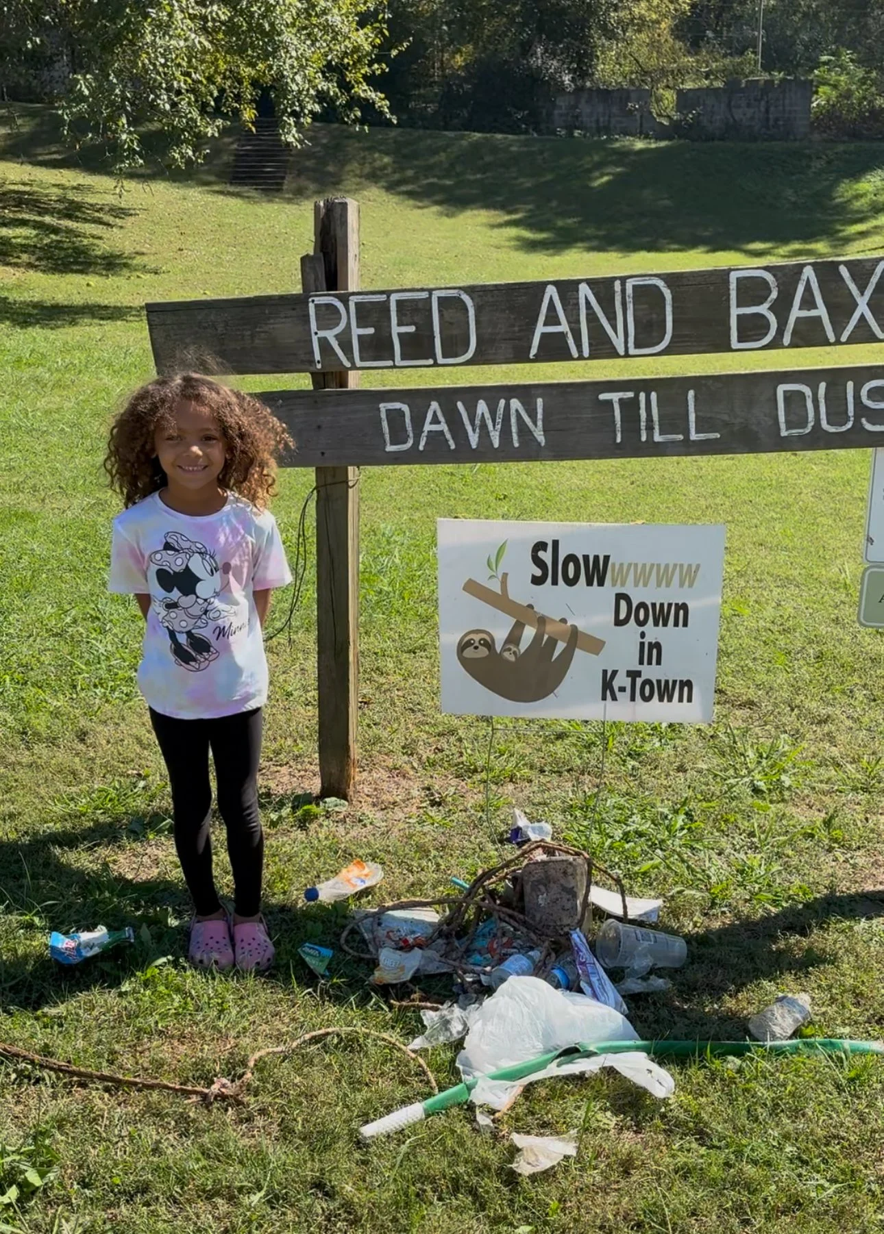 A young girl with curly hair, wearing a Minnie Mouse t-shirt, black pants, and pink Crocs, standing on a grassy area in front of a wooden sign and a smaller sign with a sloth graphic. The signs read 'REED AND BAX DAWN TILL DUS' and 'Slowww Down in K-Town.' There is trash scattered on the ground near the girl, including plastic bags, bottles, and sticks.
