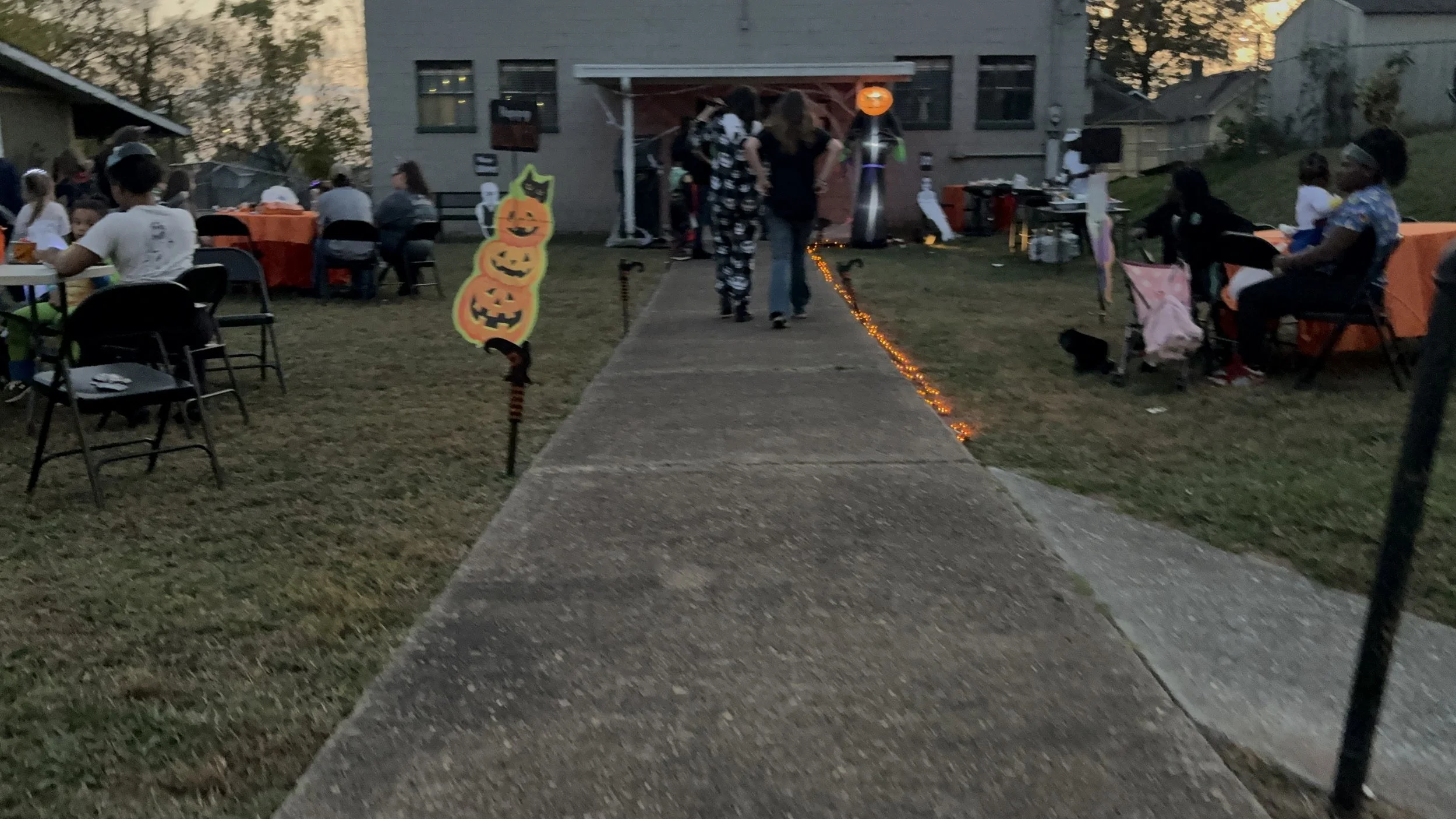People gather at a Halloween outdoor party with decorated tables, chairs, and Halloween themed decorations, including a sign with three stacked pumpkin faces and a black cat, on a residential lawn during dusk.
