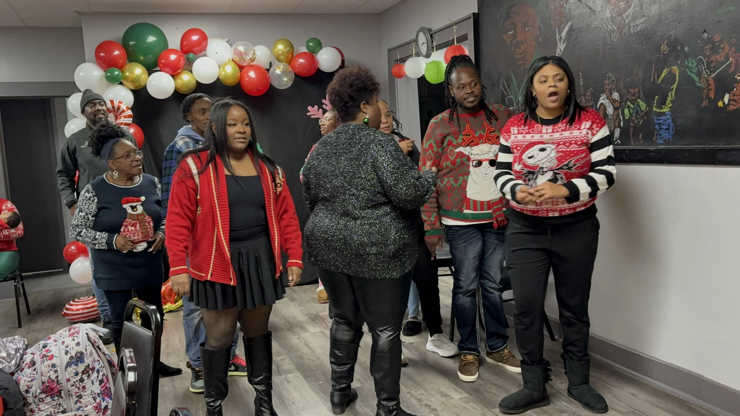 Group of people gathered indoors for a holiday celebration, wearing Christmas sweaters and festive attire, with balloons and holiday decorations in the background.