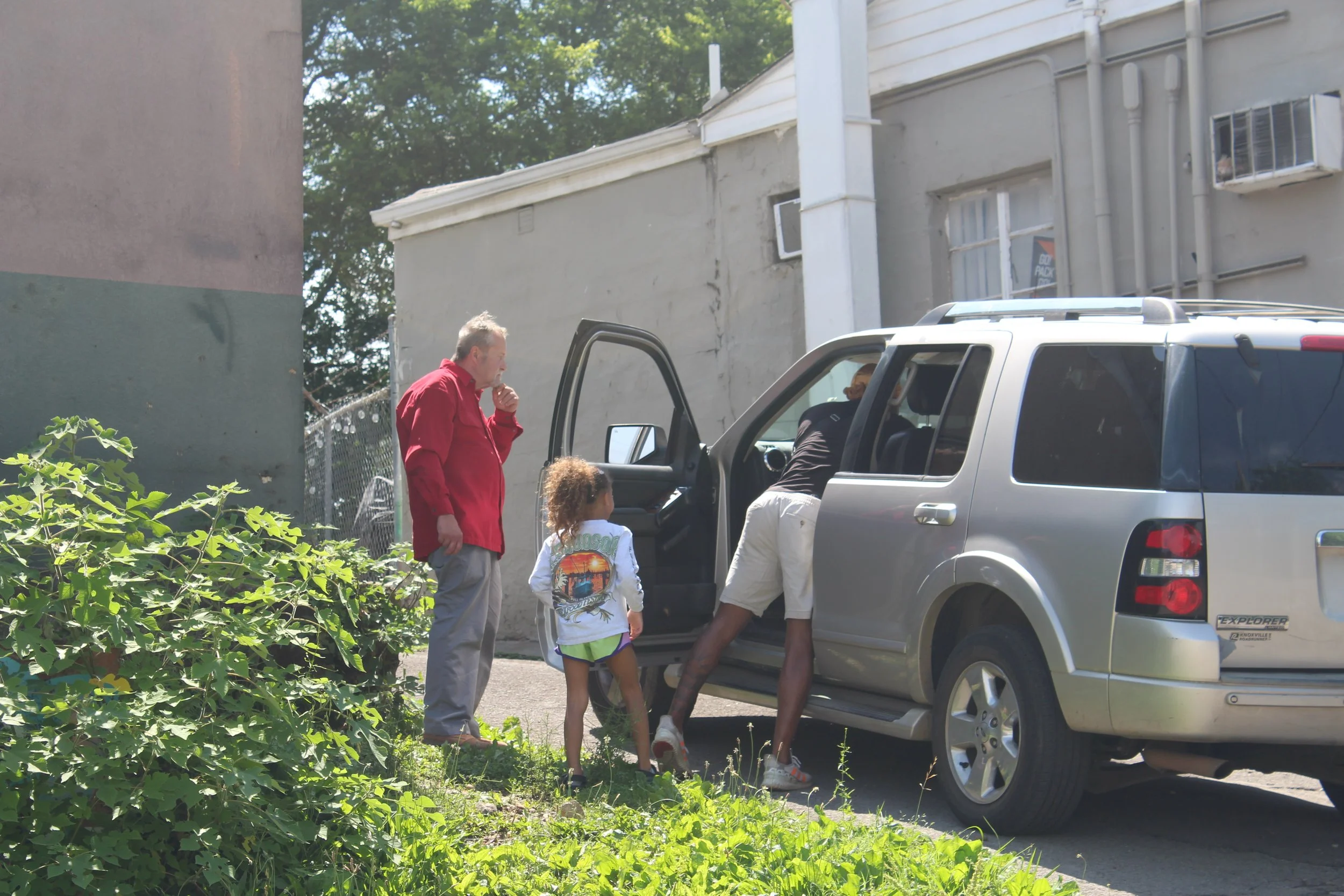 A man with gray hair in a red jacket, a young girl with curly hair and a woman with dark hair in a black shirt and white shorts, standing outside near a silver SUV, with buildings and green foliage in the background.